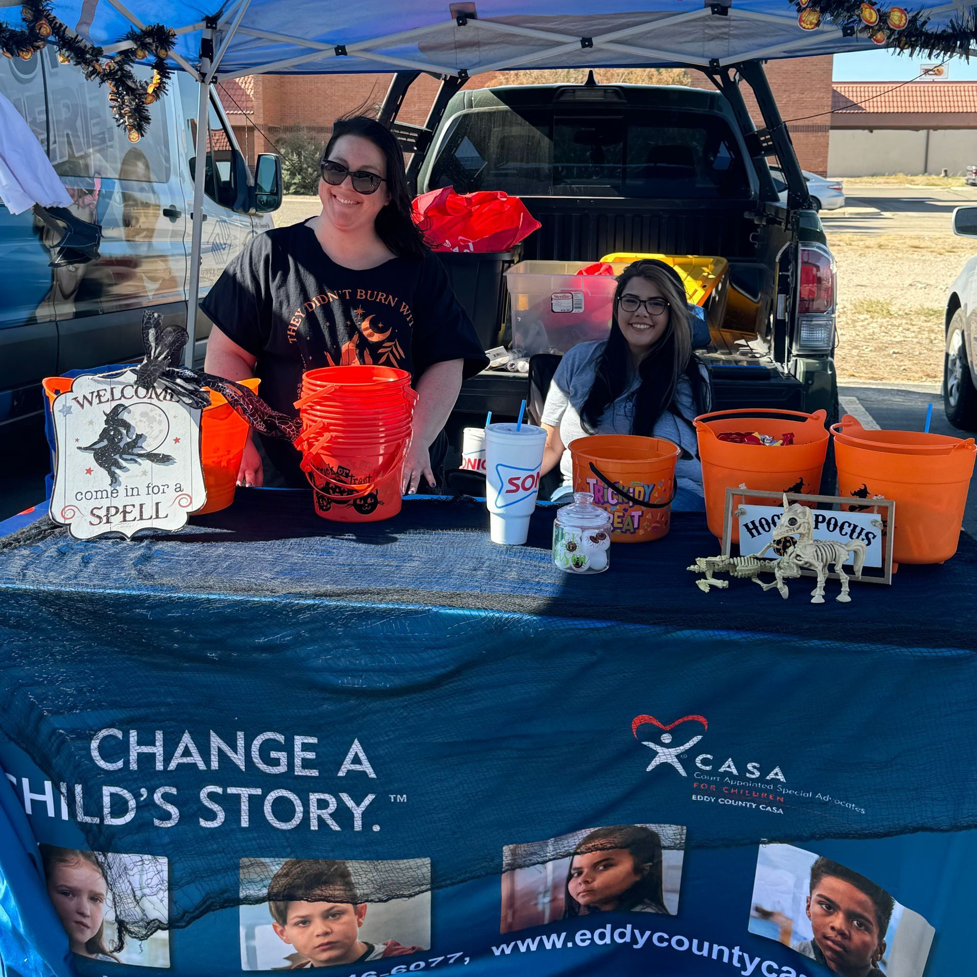 Two people at a blue-canopy booth with orange buckets and a “Change a Child’s Story” CASA banner