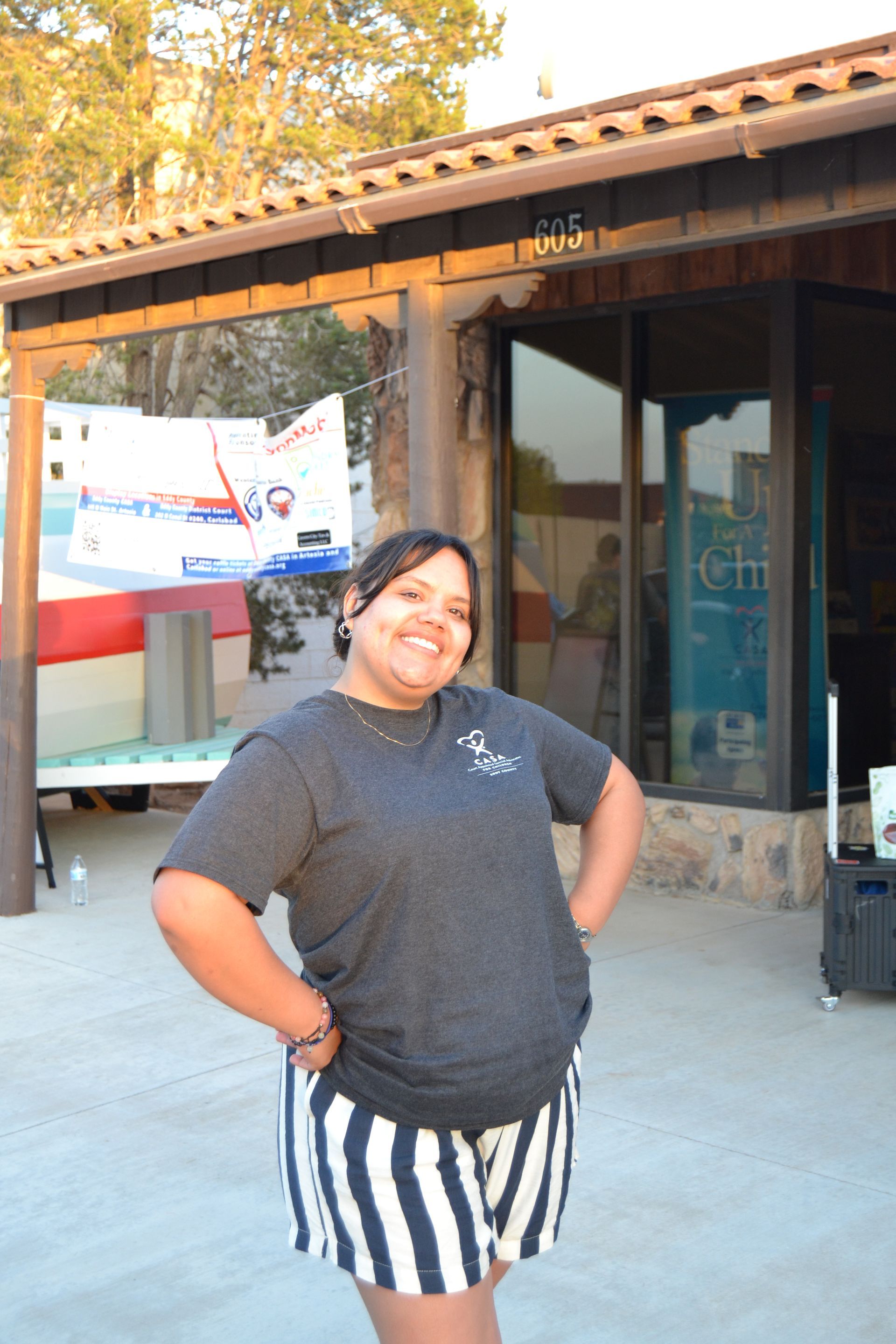 Person smiling outdoors in front of a building, wearing a dark shirt and striped shorts.
