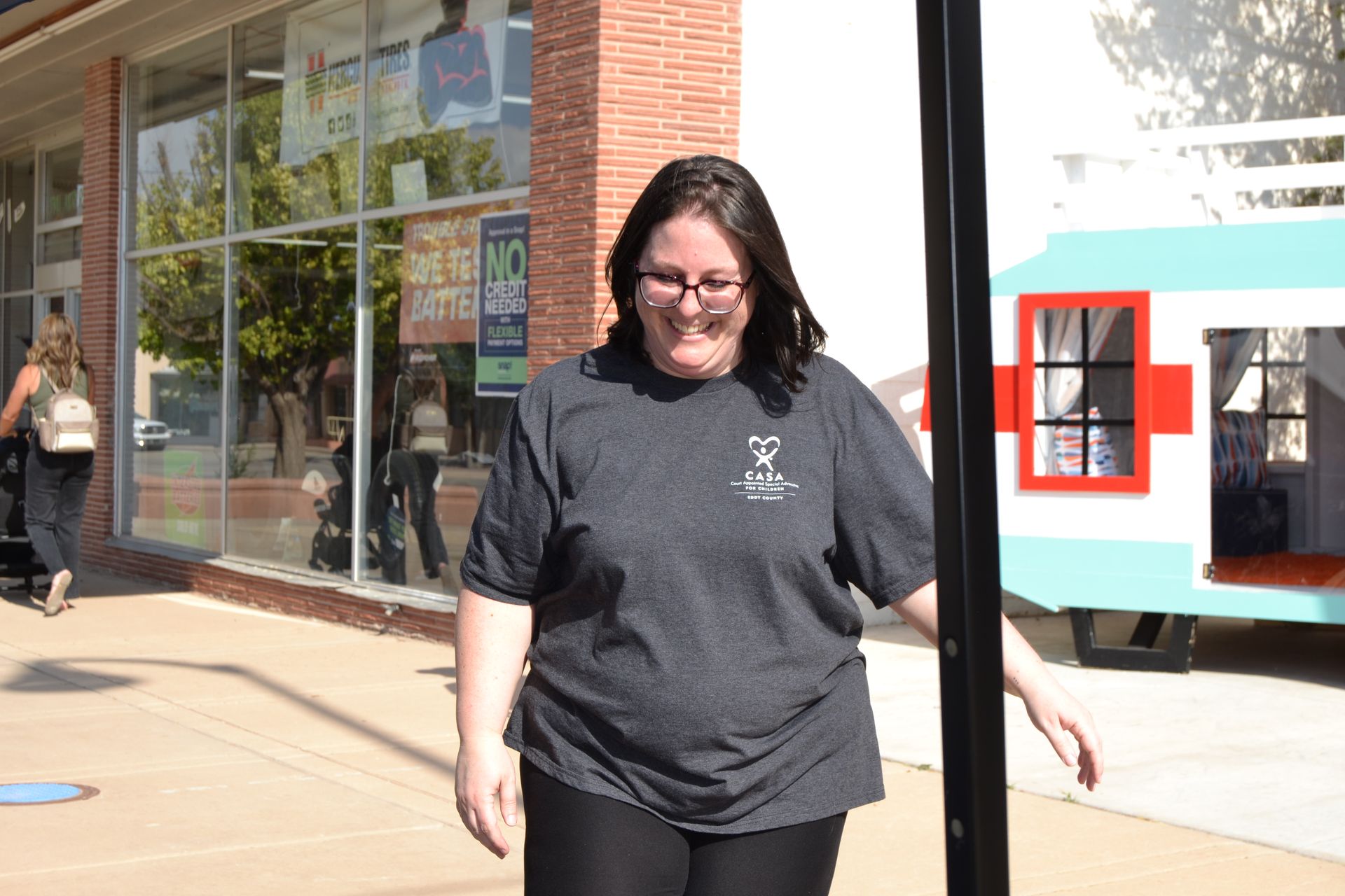 Person walking outside a storefront, smiling, in a black T-shirt and glasses