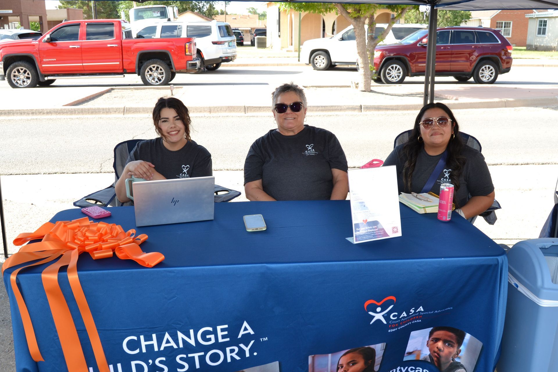 Three people at a blue event table with laptops and signs, under a tent near a street.