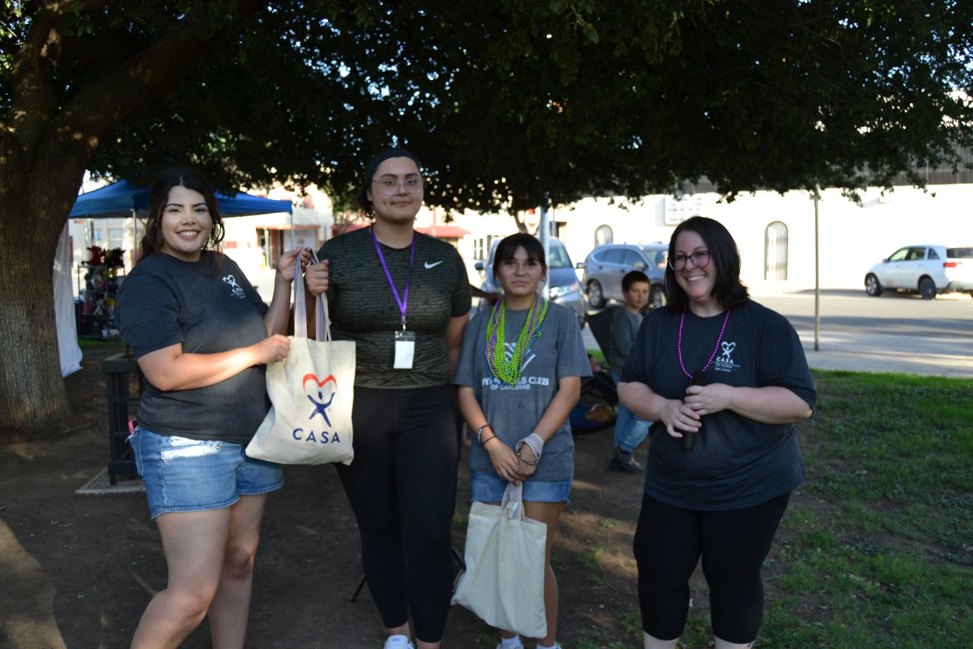 Four people standing outdoors under a tree, holding bags and smiling at the camera.
