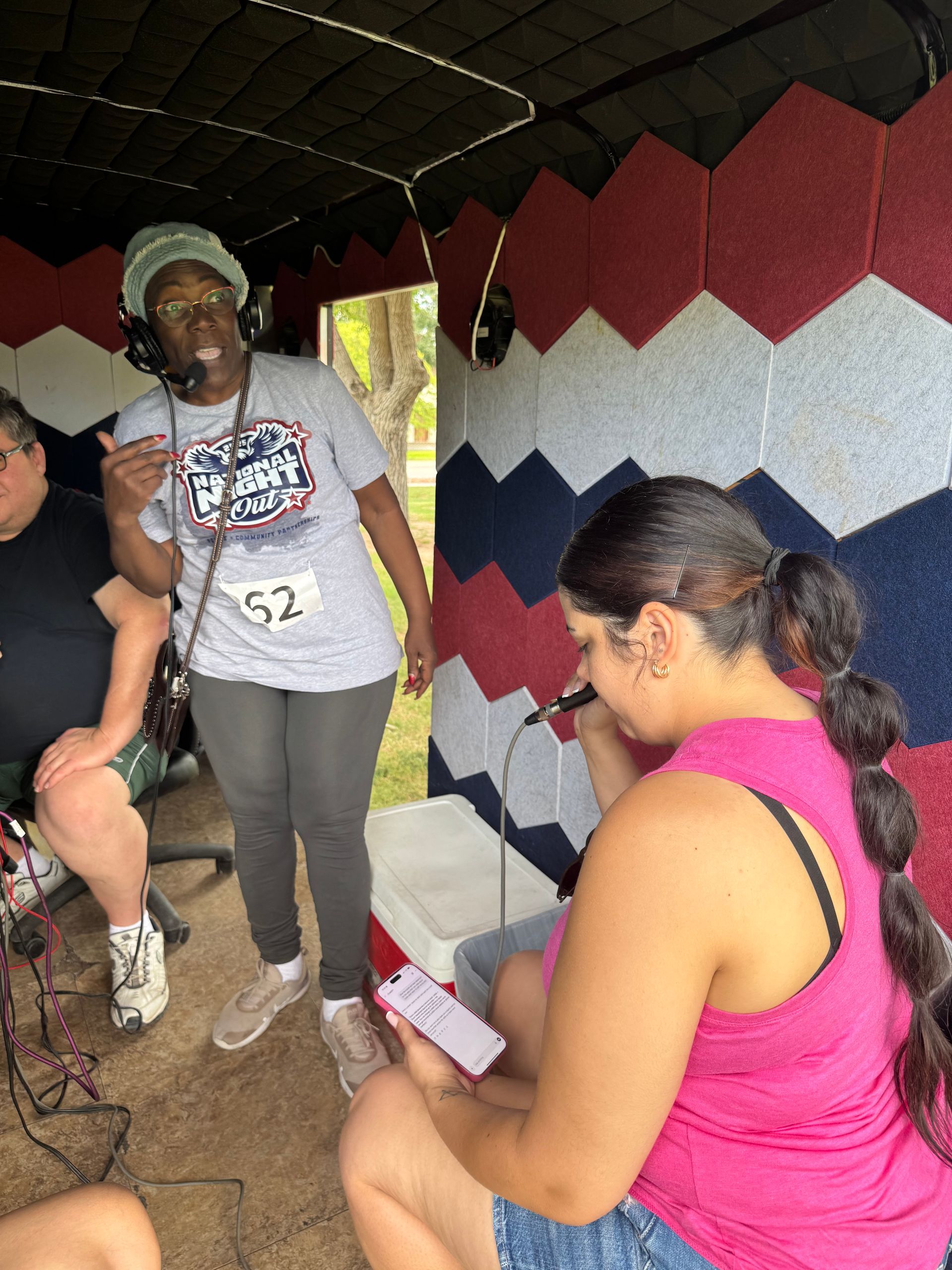 Three people under a canopy, one speaking into a microphone beside a red, white, and blue quilted wall.