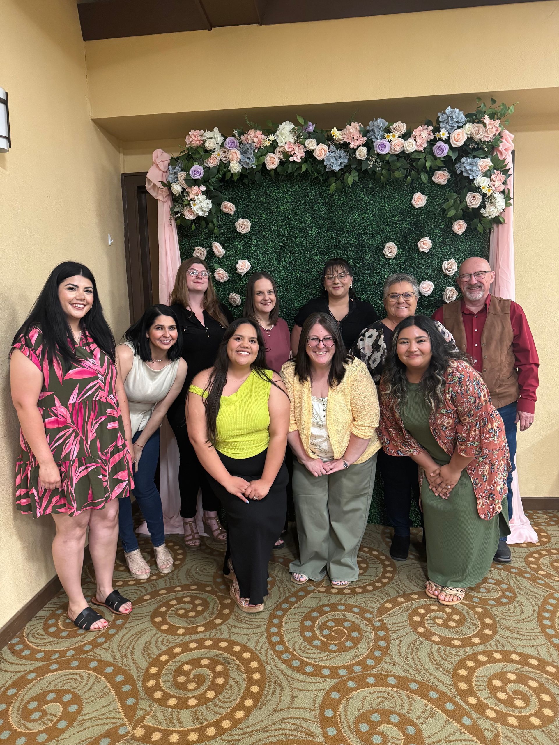 Group posing indoors before a floral backdrop, smiling and dressed in colorful outfits