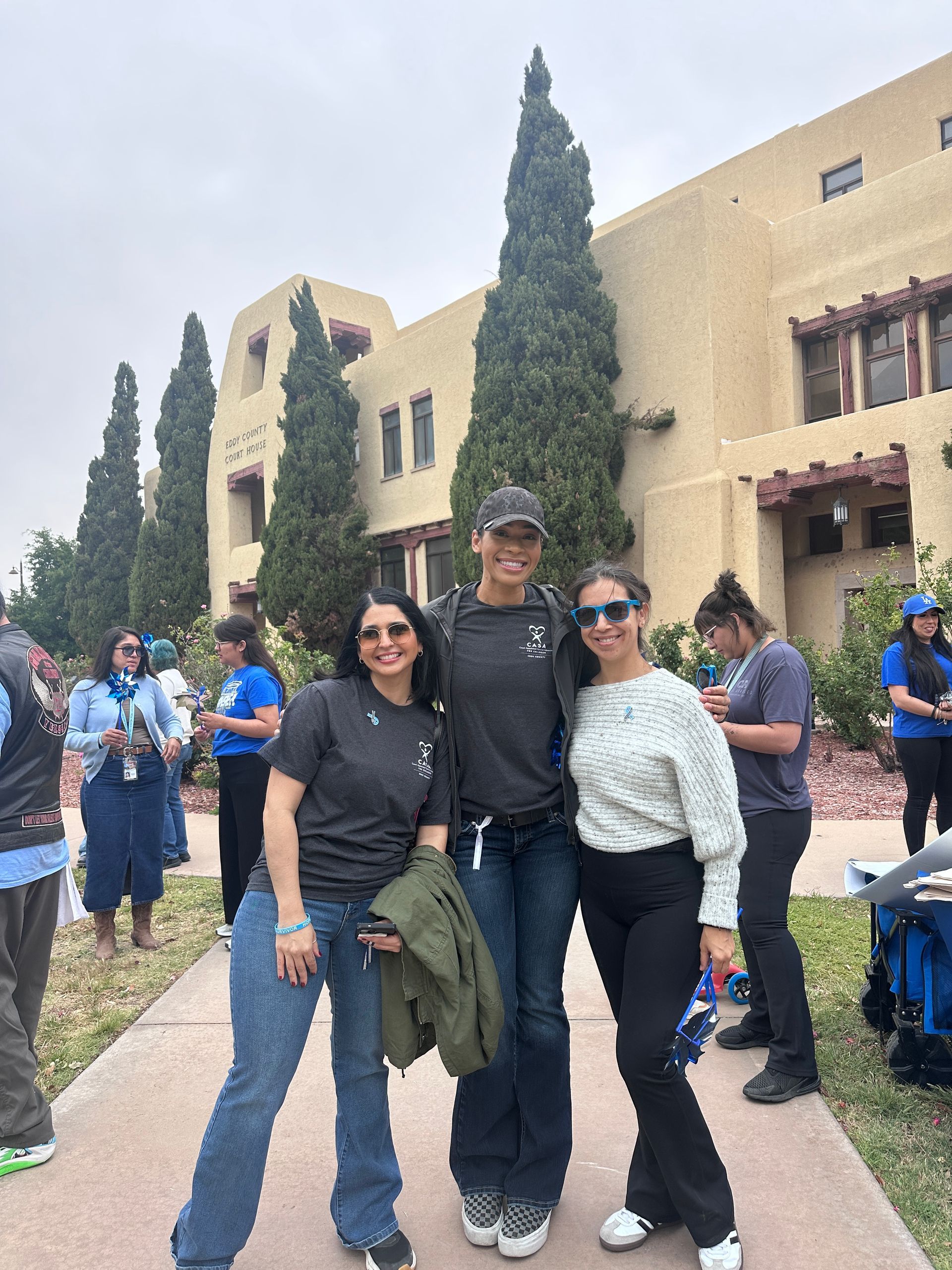 Three people posing outdoors on a campus walkway beside a tan building and tall trees