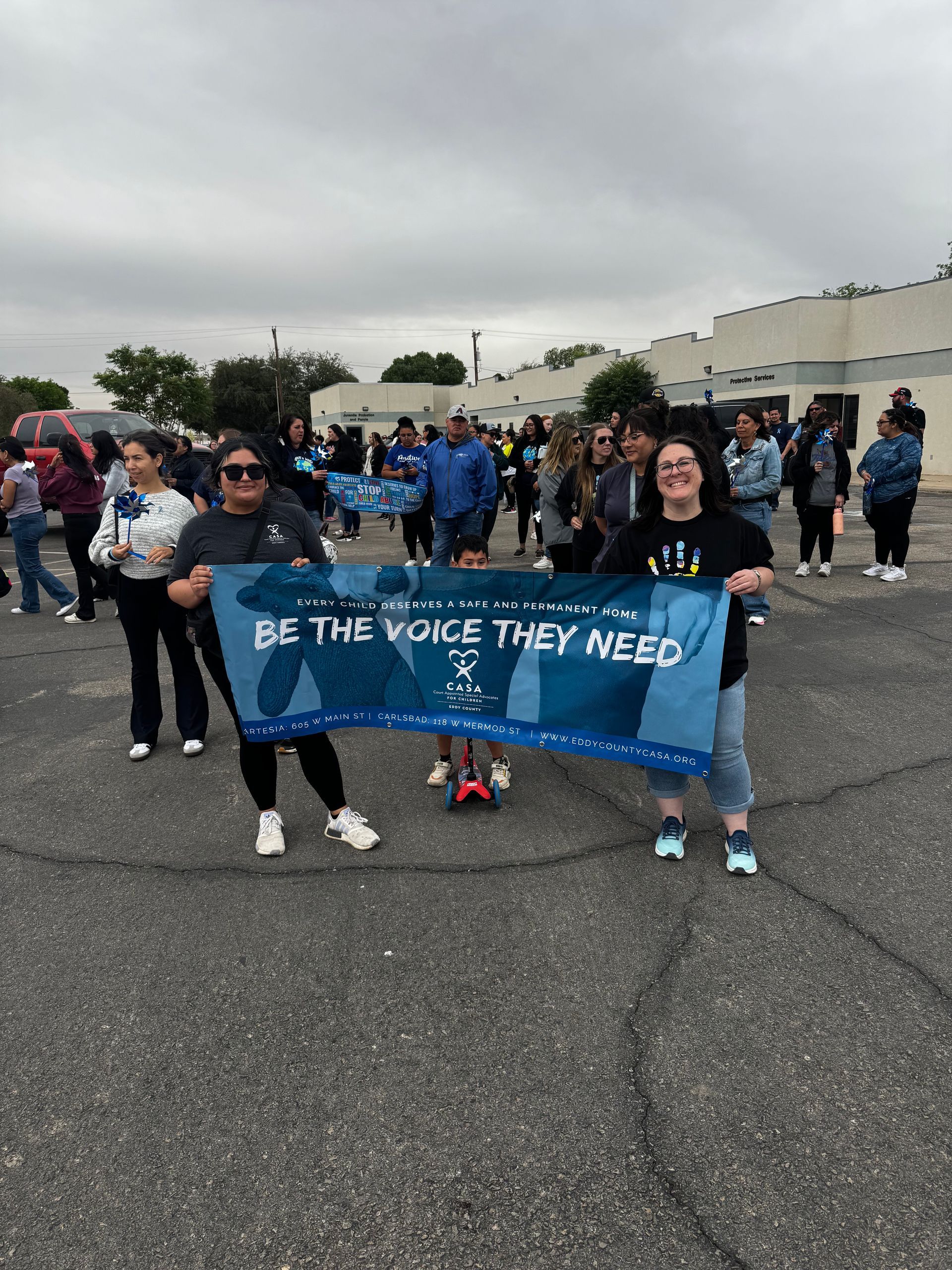 Crowd holding a blue banner during a march in a parking lot under cloudy skies