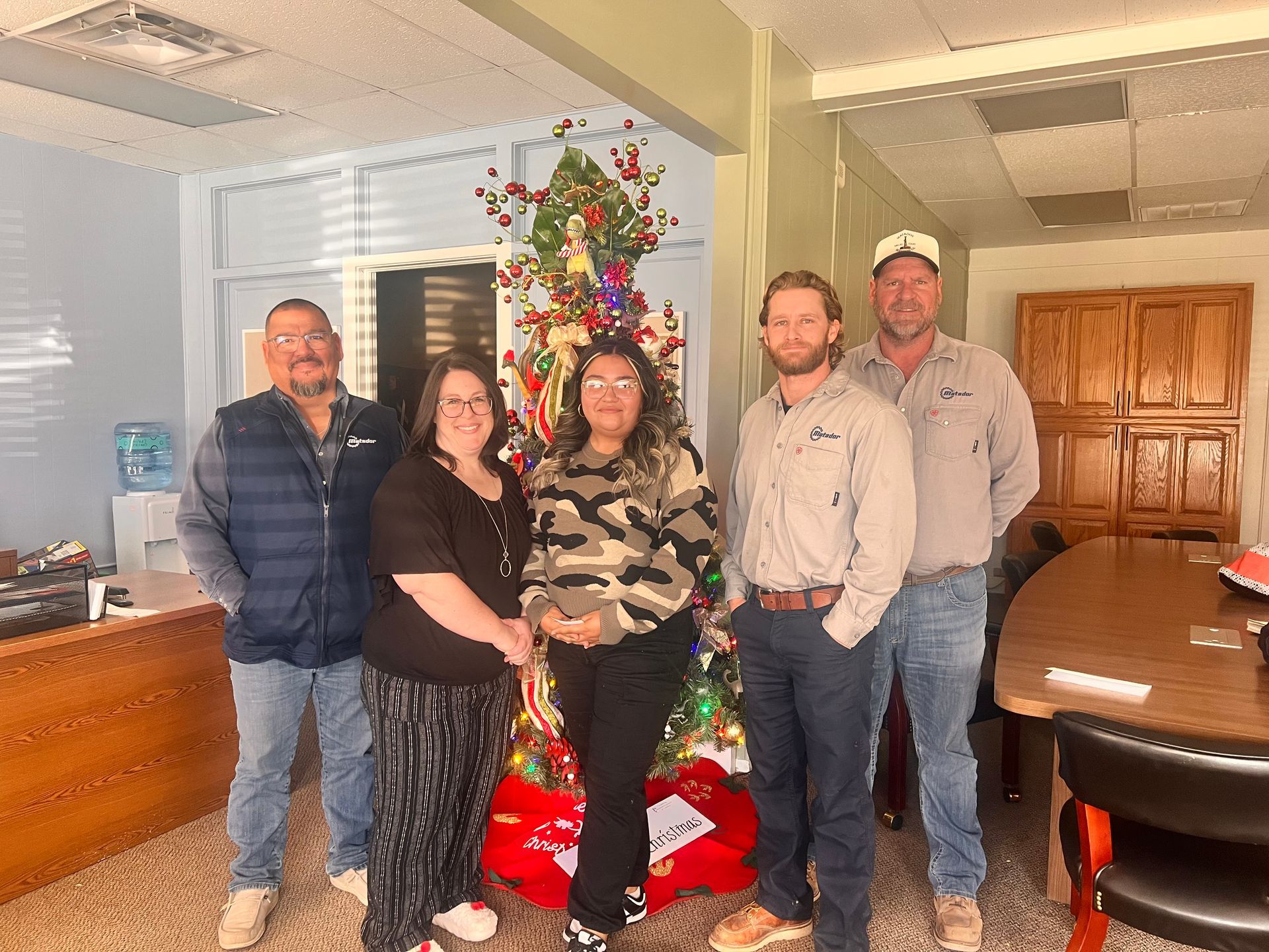 Five people posing in an office beside a decorated Christmas tree and wrapped gifts