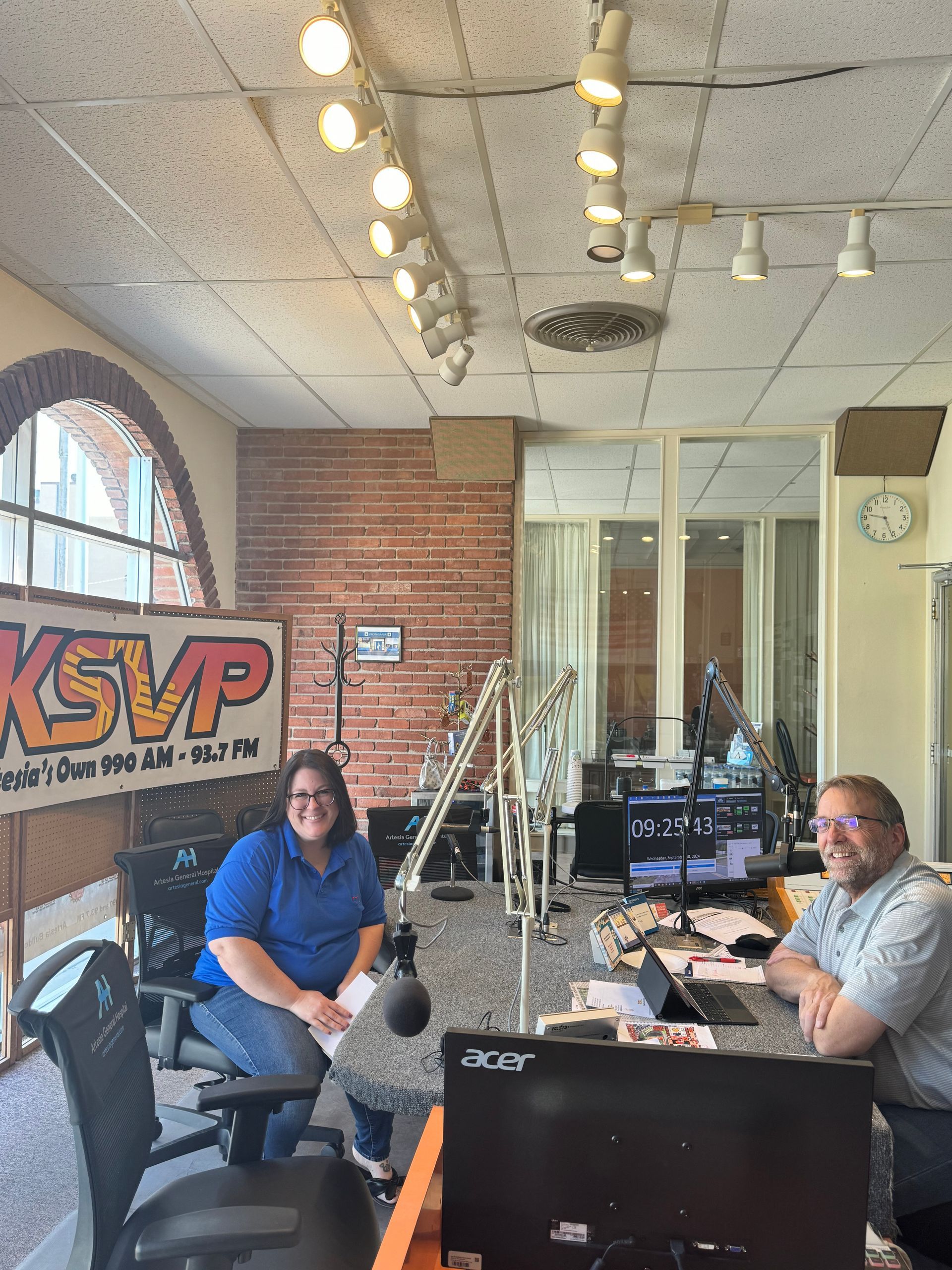 Two people seated at a radio station studio with microphones, mixer, and a KSVP sign in the background