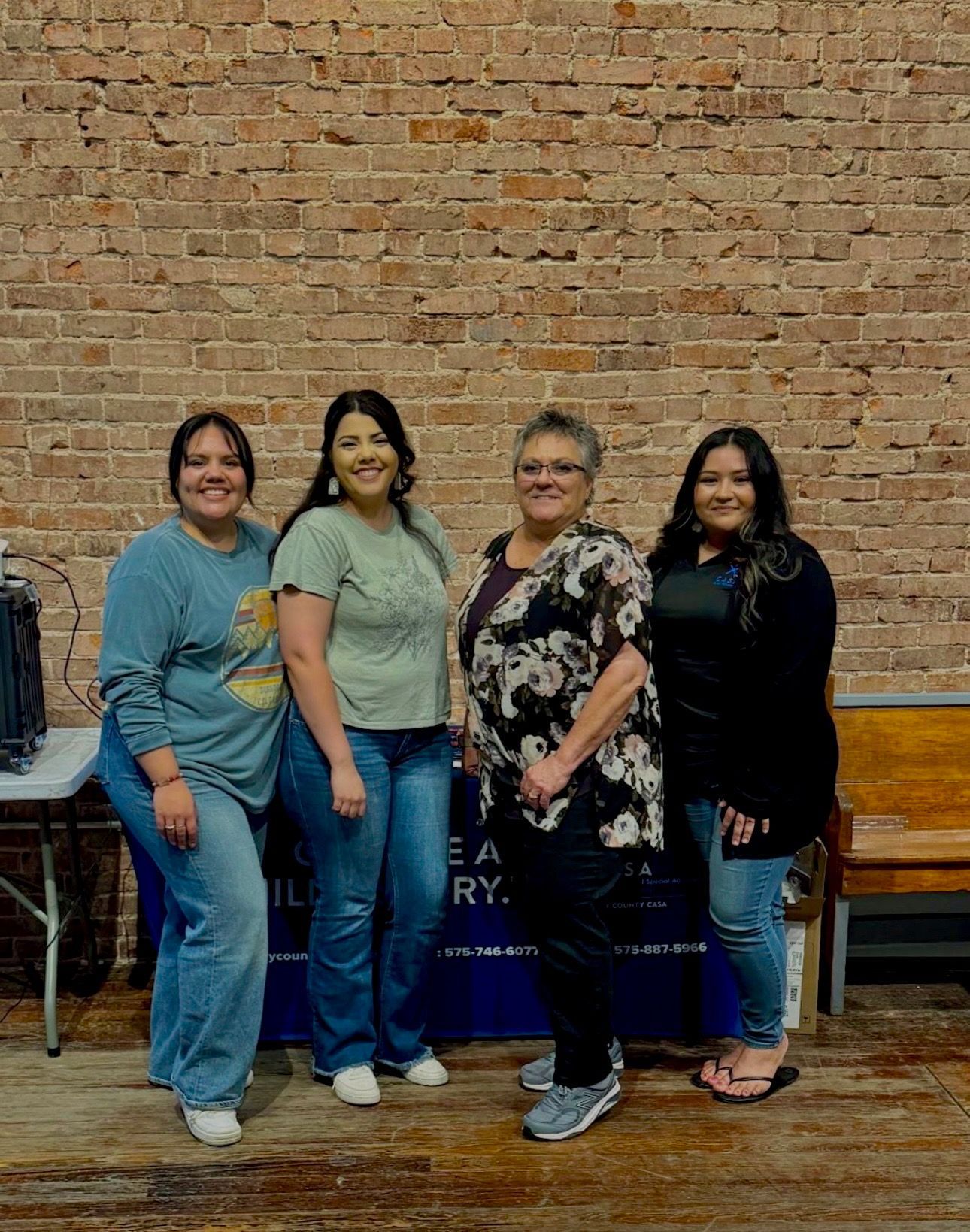 Four people posing together indoors against a brick wall, smiling and standing side by side.
