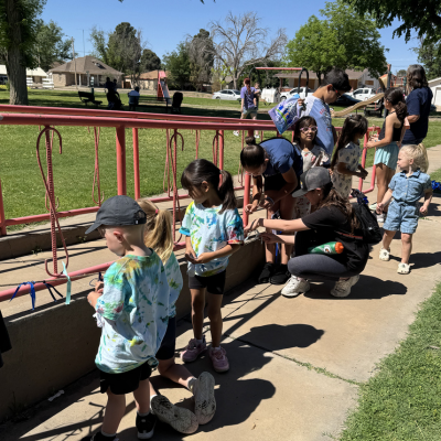 Children stand in line outdoors while a crouching adult helps one child near red railings on a sunny day