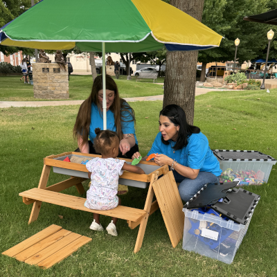 Two adults and a child at a wooden picnic table under a green-and-yellow umbrella outdoors