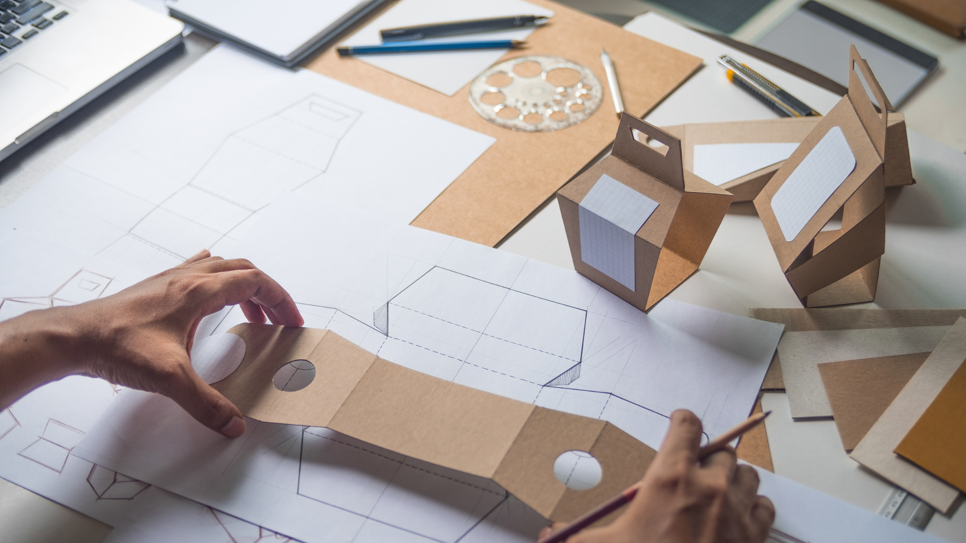 Des mains dessinent des maquettes d'emballage avec du matériel en carton sur un bureau.