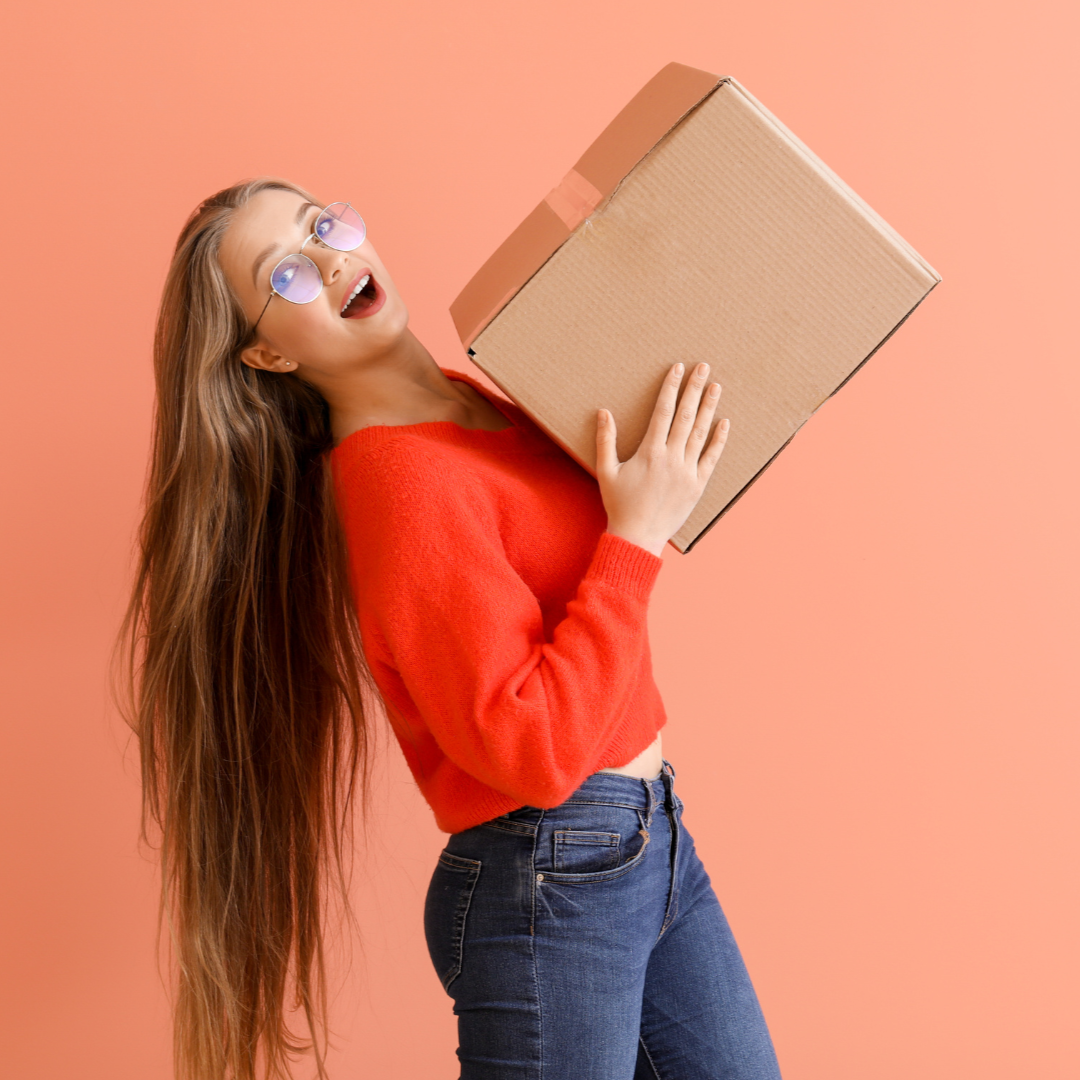 Femme en pull rouge et jean, tenant une boîte en carton, l'air enthousiaste sur fond orange.