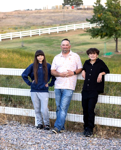A man and two young people lean against a white farm fence in a grassy field with a hillside in the background.
