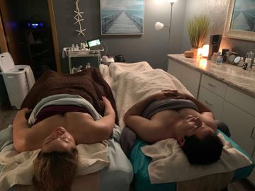 Two people lying face down on massage tables in a softly lit spa room, with candles and towels nearby