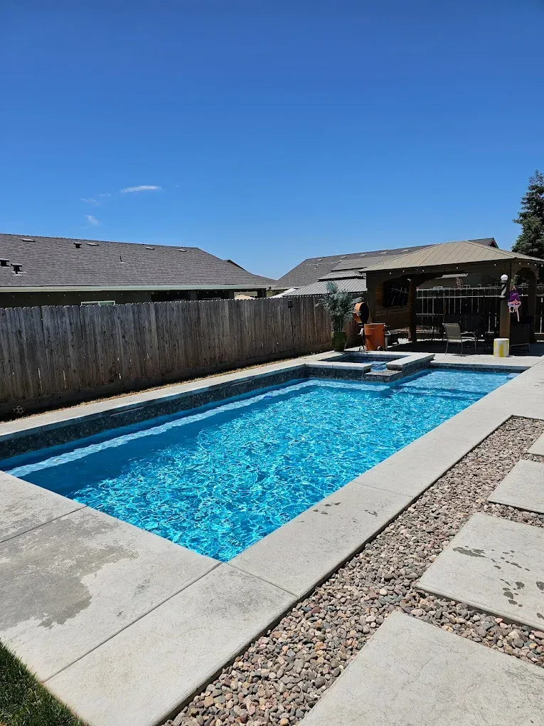 Rectangular pool with blue water. Concrete patio with stepping stones and gravel. Wooden fence and gazebo in background.