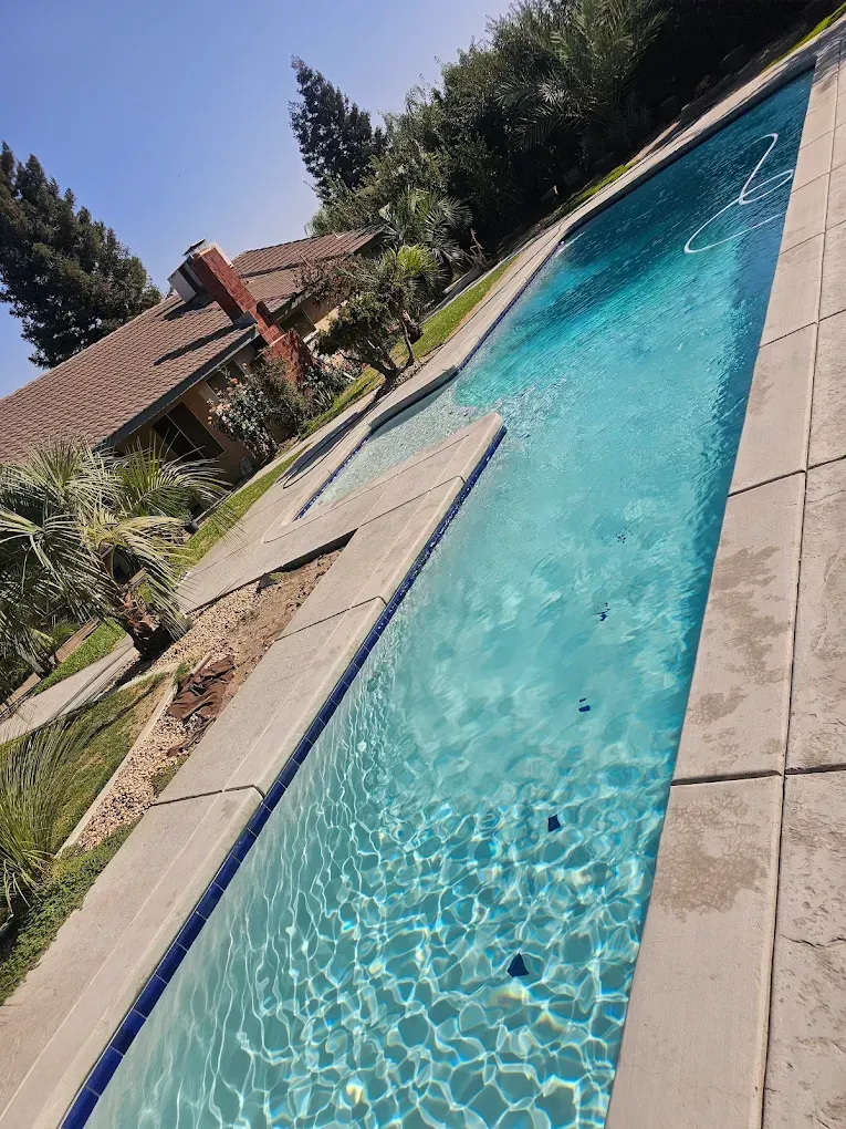Long rectangular swimming pool, turquoise water, concrete surround, beside grass and house under a blue sky.