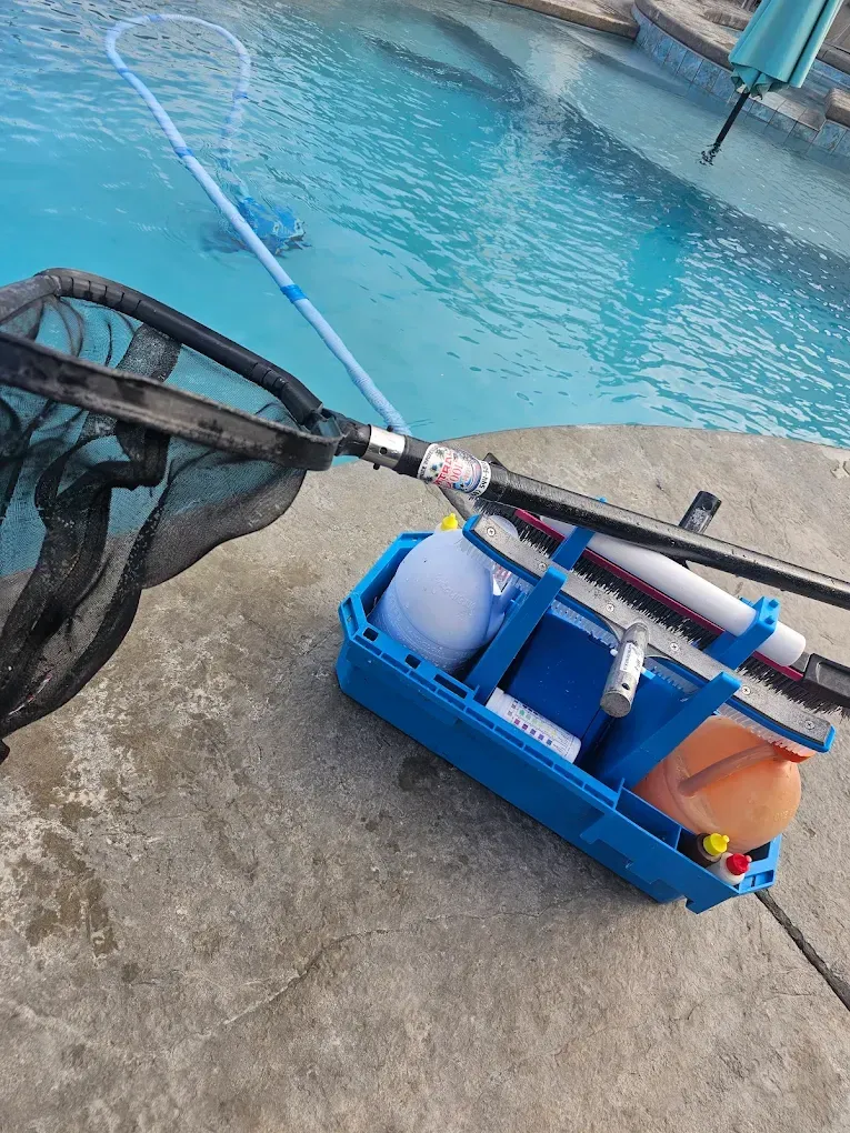 Pool cleaning supplies on a concrete deck. Blue net and vacuum, container holds supplies near a pool.