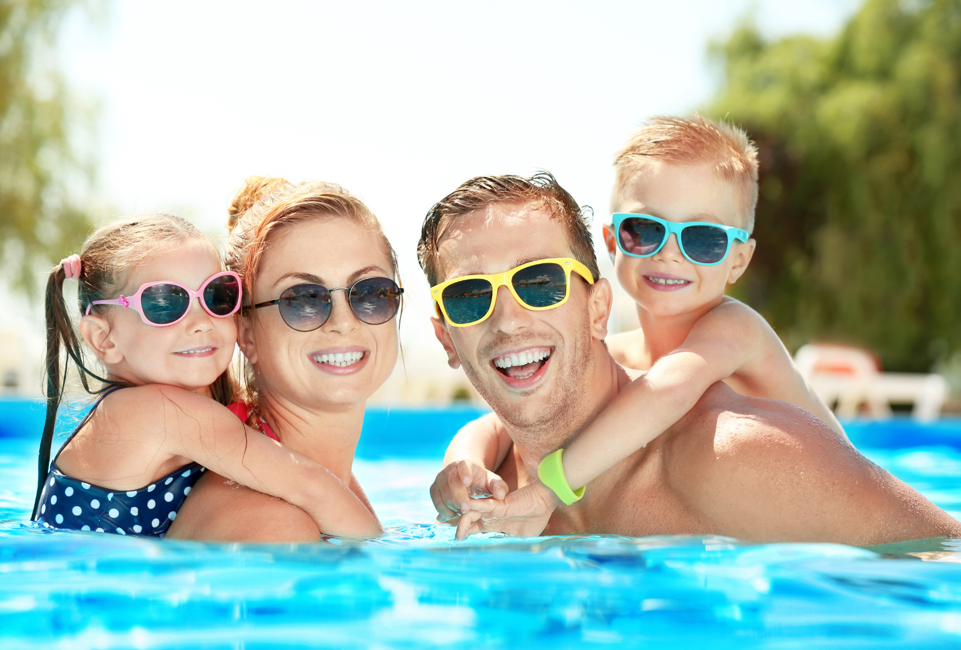 Family smiling in a pool, wearing sunglasses. Two children on their parents’ backs. Blue water, sunny setting.
