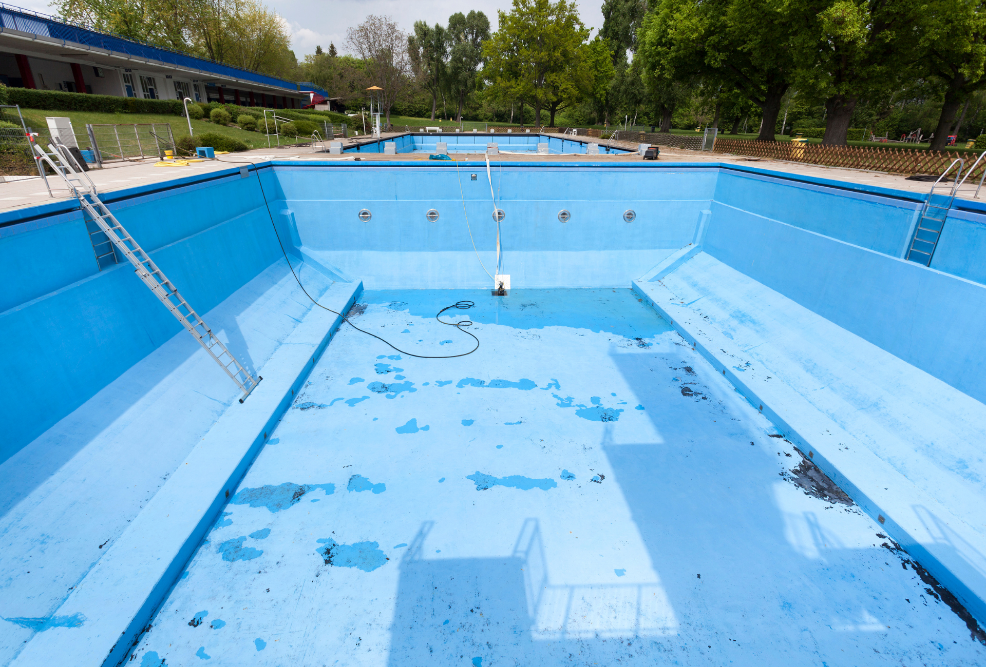Empty blue swimming pool, concrete surrounds. Ladders lead into the empty basin. Green trees and buildings in background.