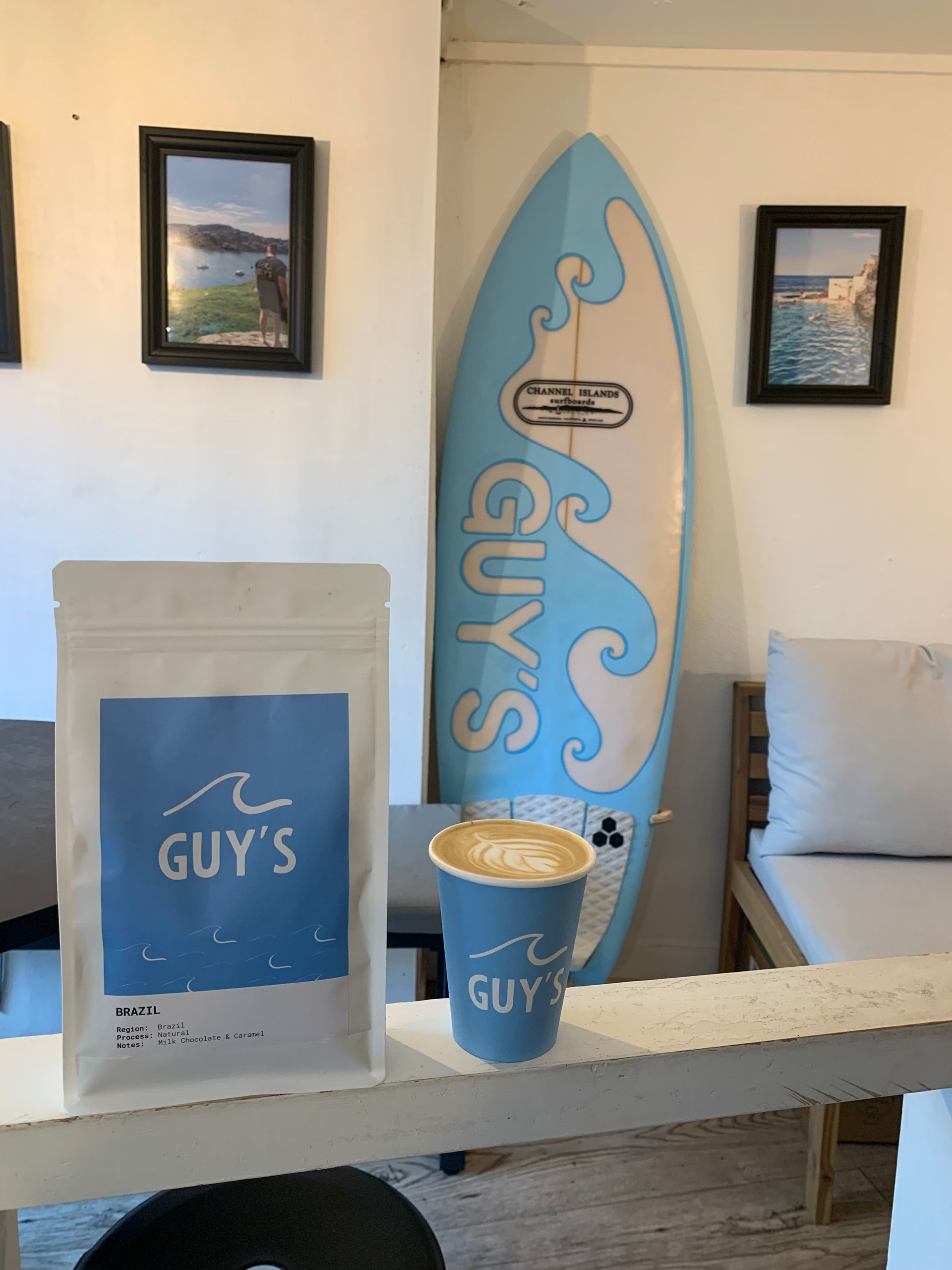 Coffee shop scene: Coffee bag, latte, blue surfboard, photos on wall, white interior.