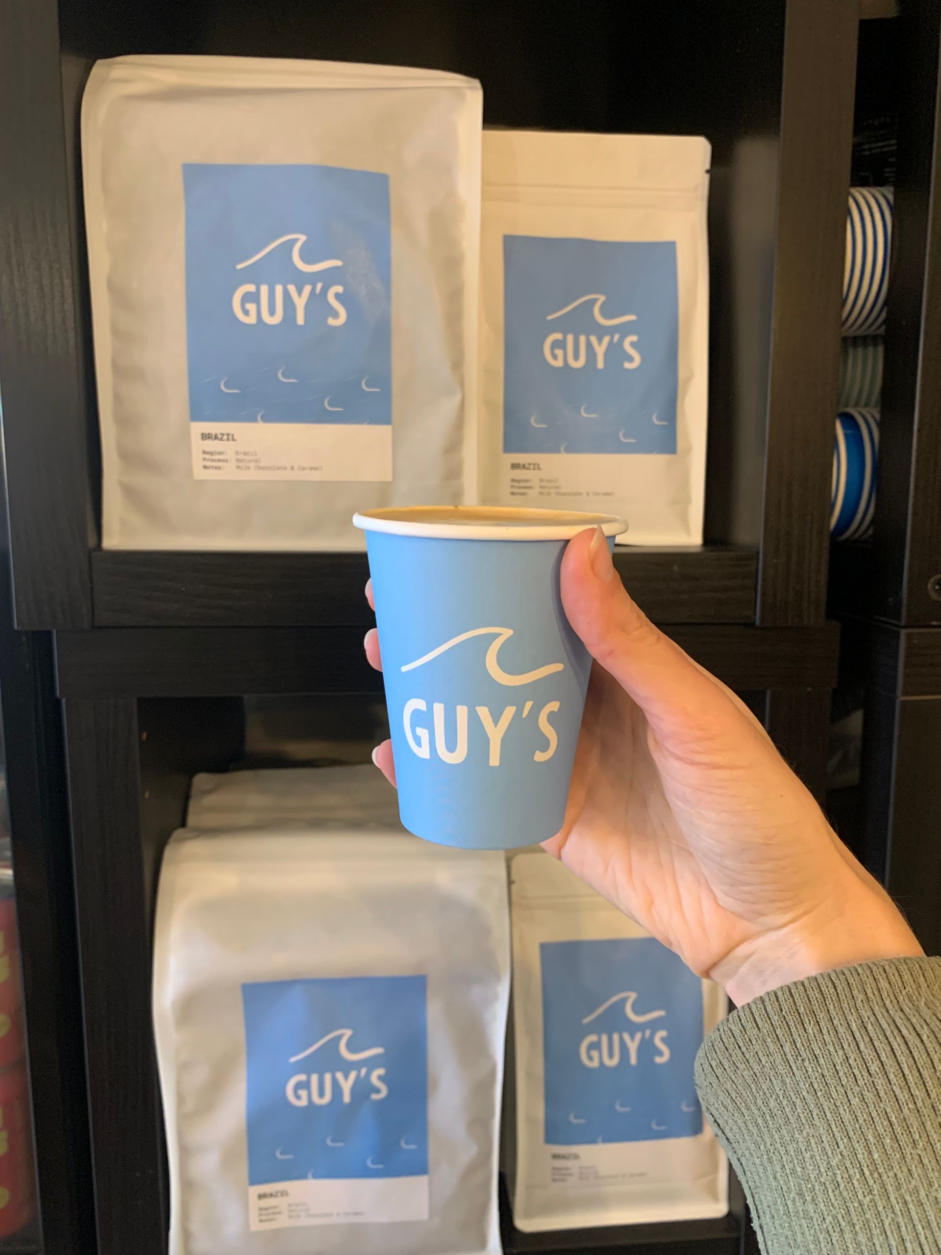 Coffee shop scene: Coffee bag, latte, blue surfboard, photos on wall, white interior.