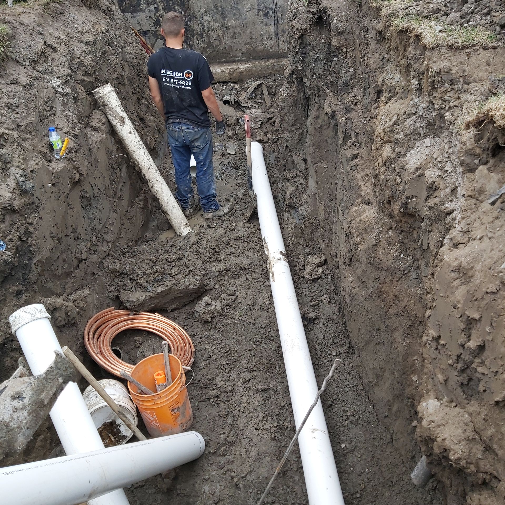 Un homme se tient debout dans une tranchée boueuse avec des tuyaux et un seau.
