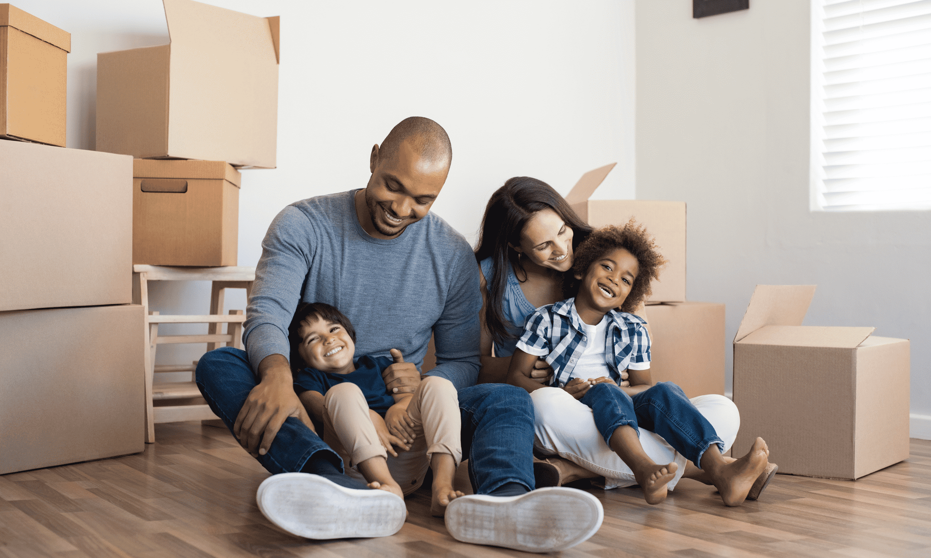 A family is sitting on the floor in a room filled with cardboard boxes.