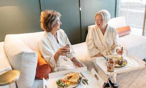 Two women are sitting on a couch eating food and drinking wine.