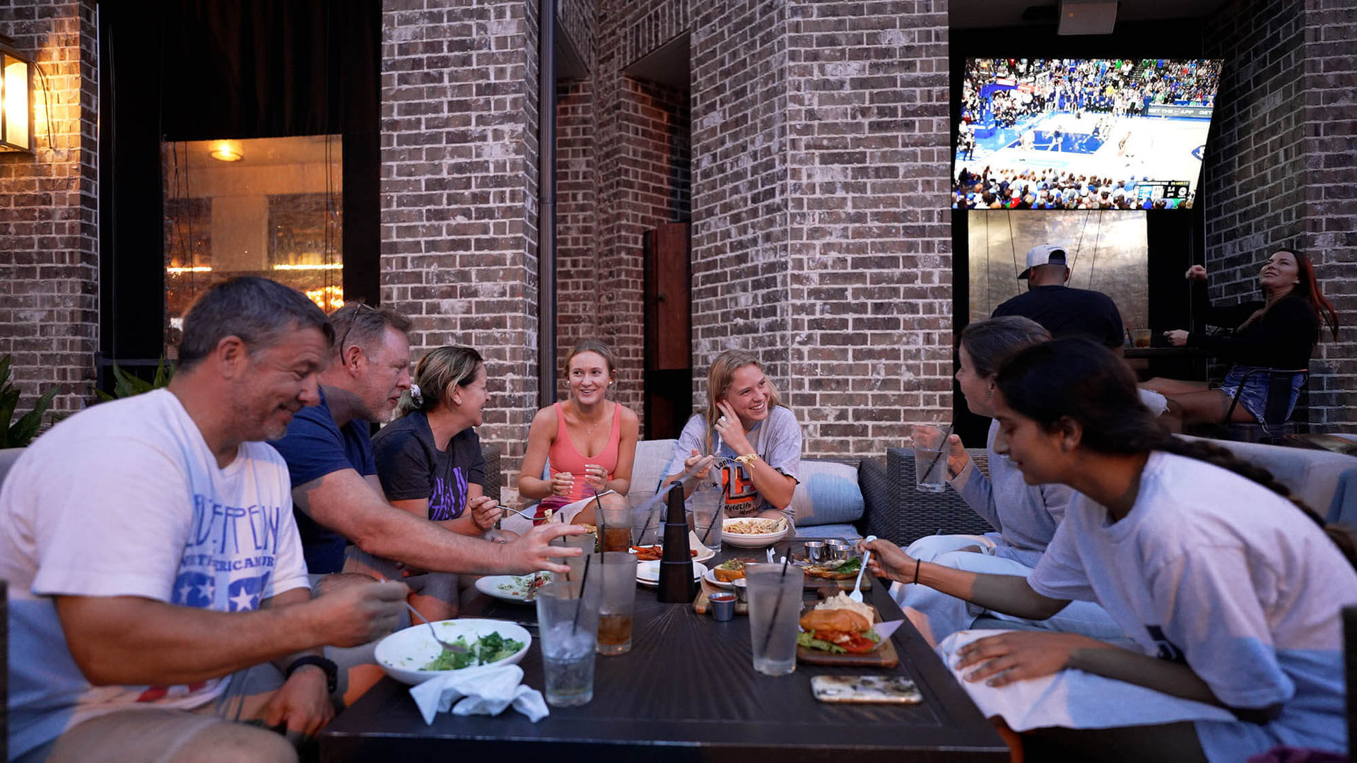 People sitting at an outdoor table, laughing and eating. Brick wall background, TV screen playing a game.