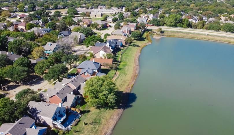 Aerial view of a residential area with houses lining a lake, green trees, and a cloudy blue sky.