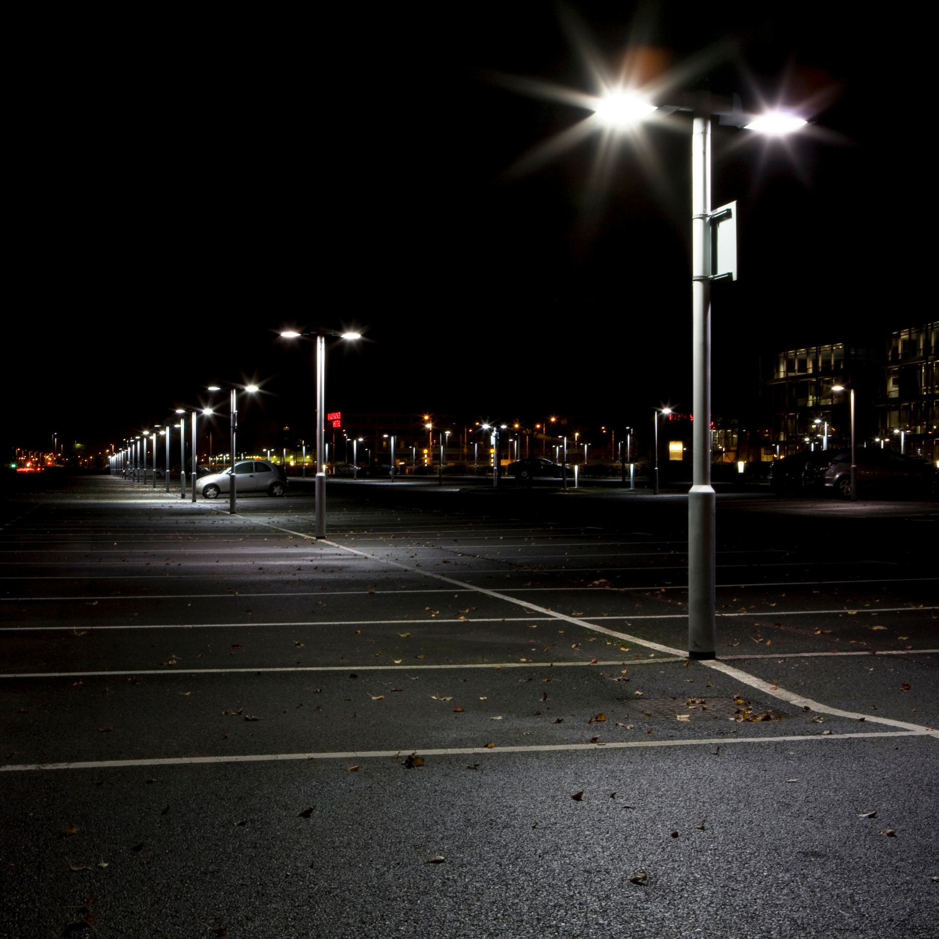Empty parking lot at night, lit by bright streetlights, with city lights in the distance.