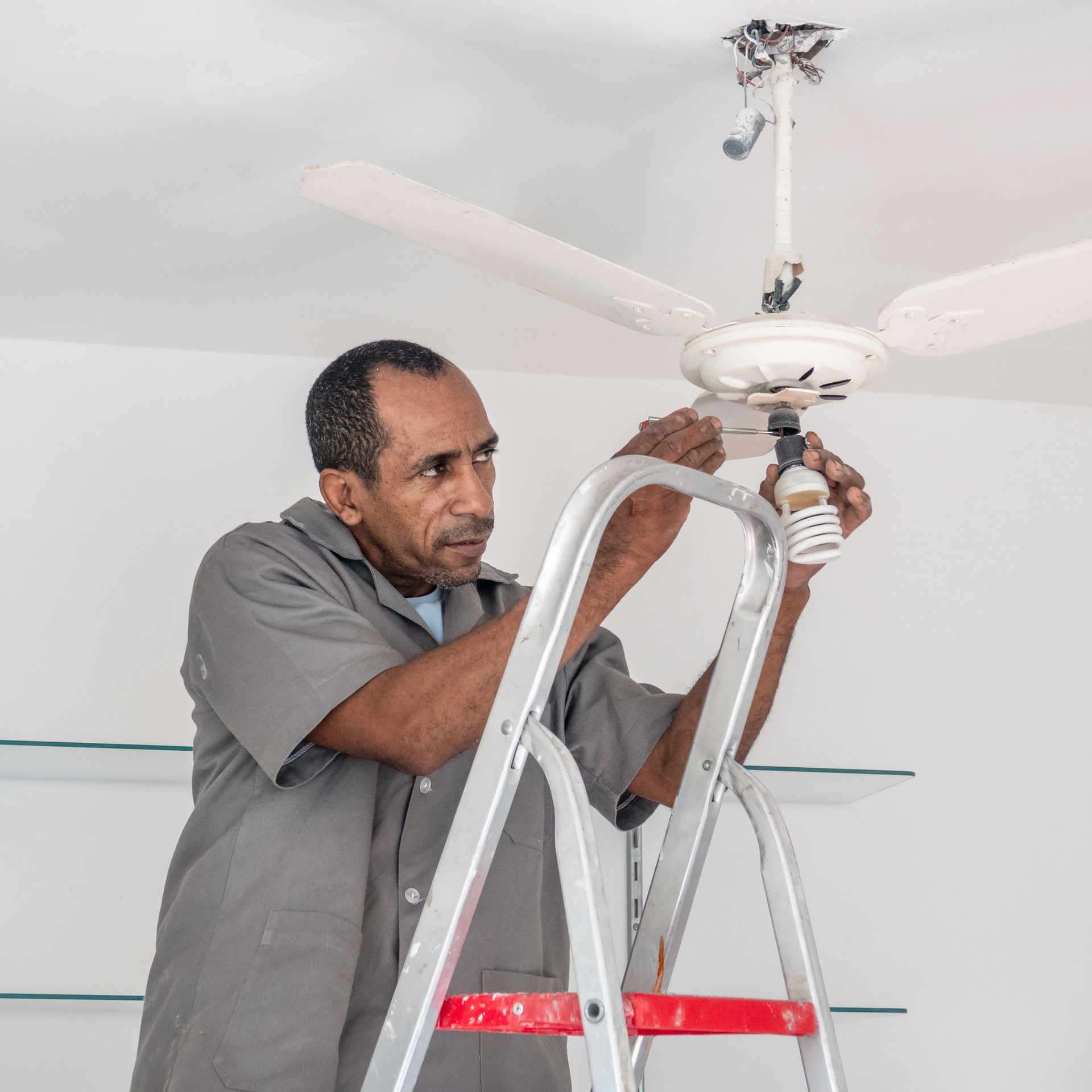 Man on a ladder changing a lightbulb in a ceiling fan. White walls, gray uniform.