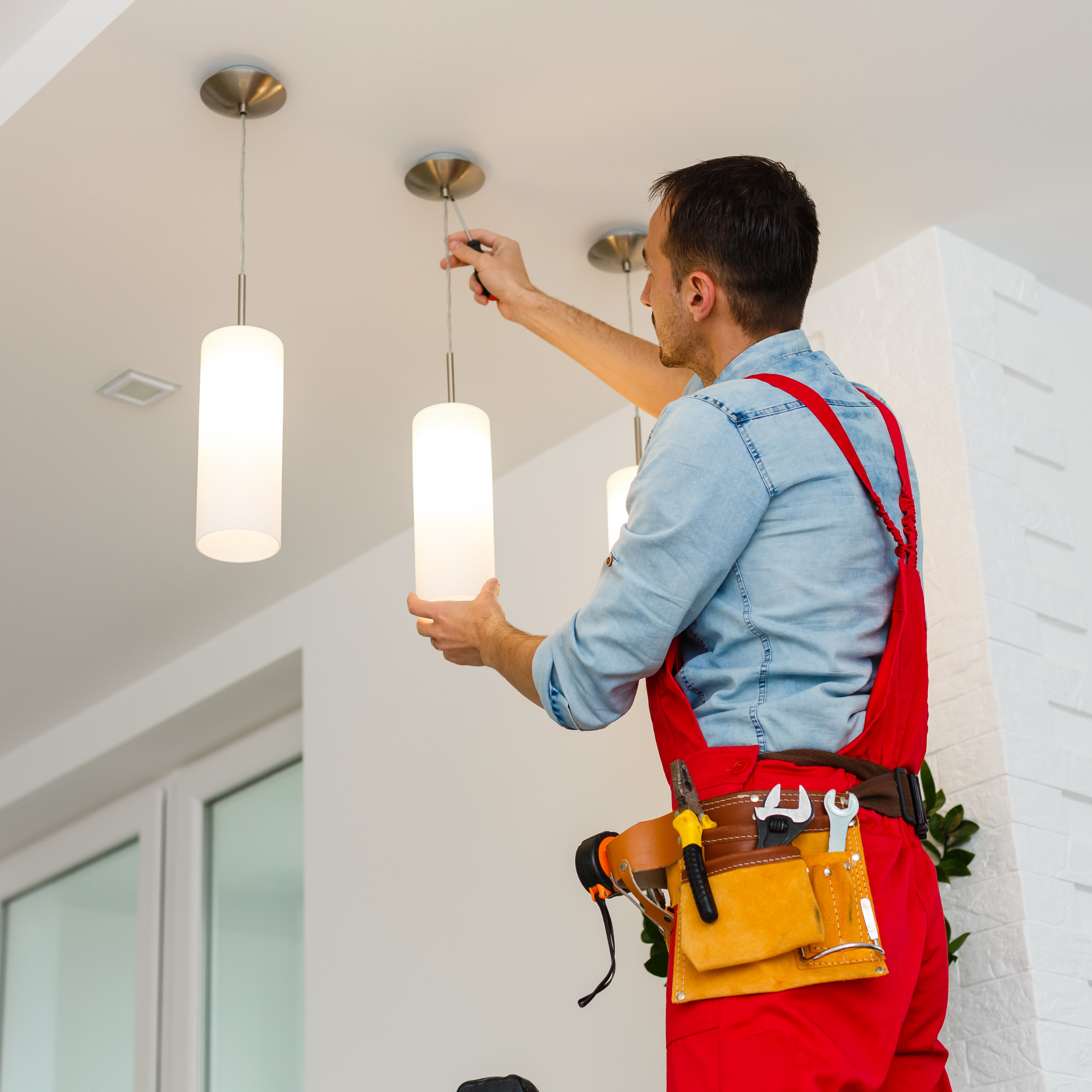 Man in red overalls installing a pendant light fixture on a white ceiling in a home.