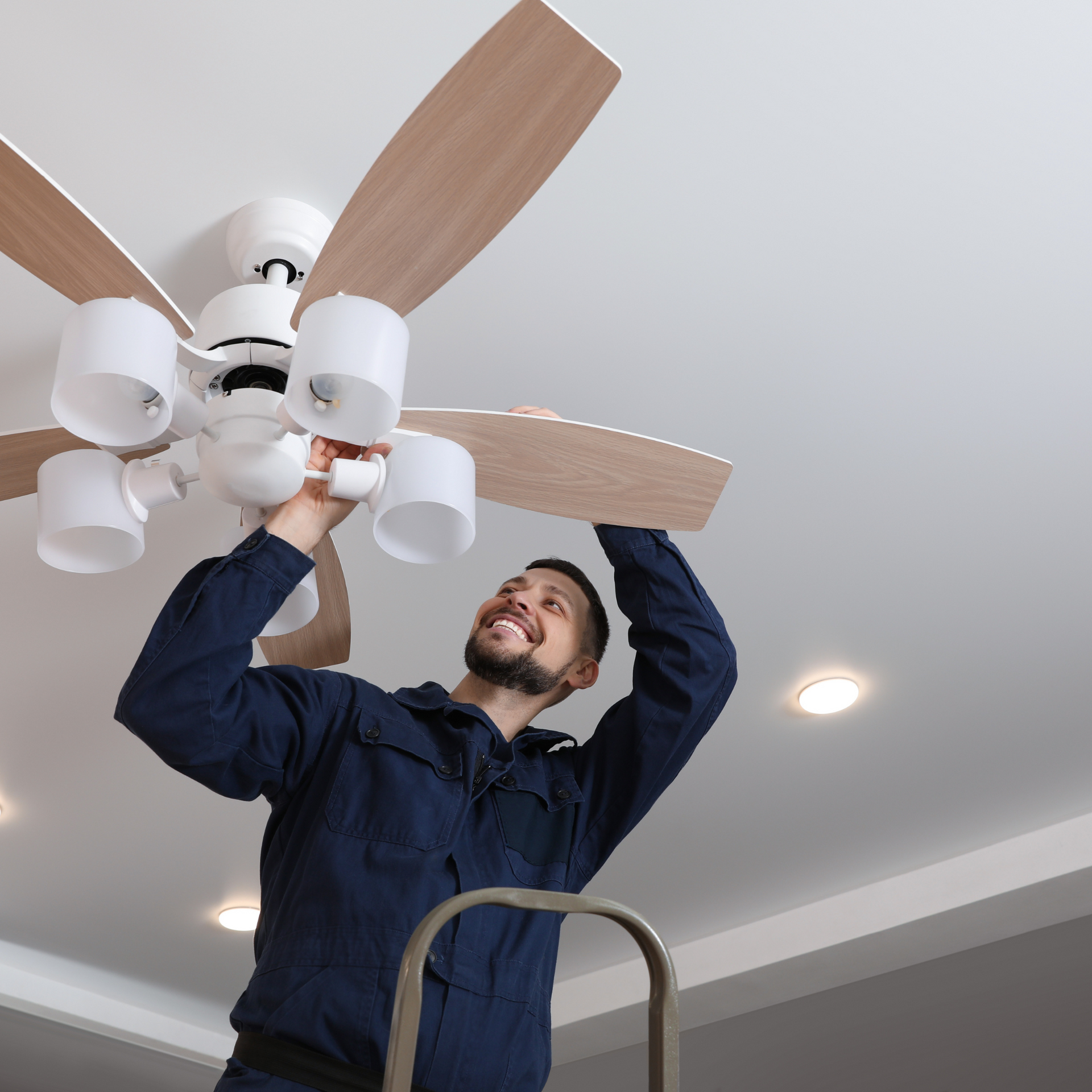 Man installing ceiling fan, smiling. He wears blue work suit, stands on ladder, bright room.