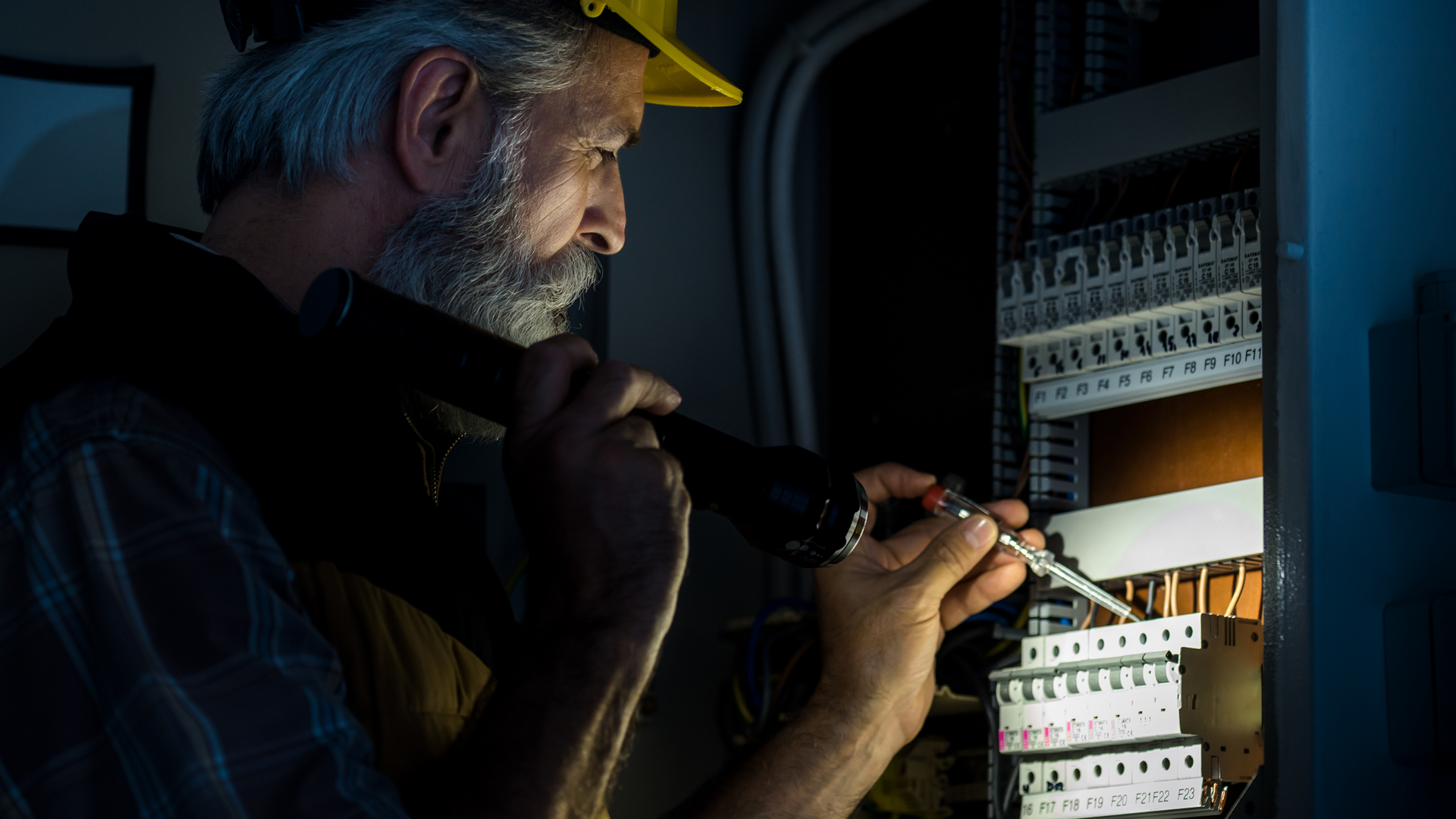 Electrician with a flashlight working on an electrical panel in a dim room.