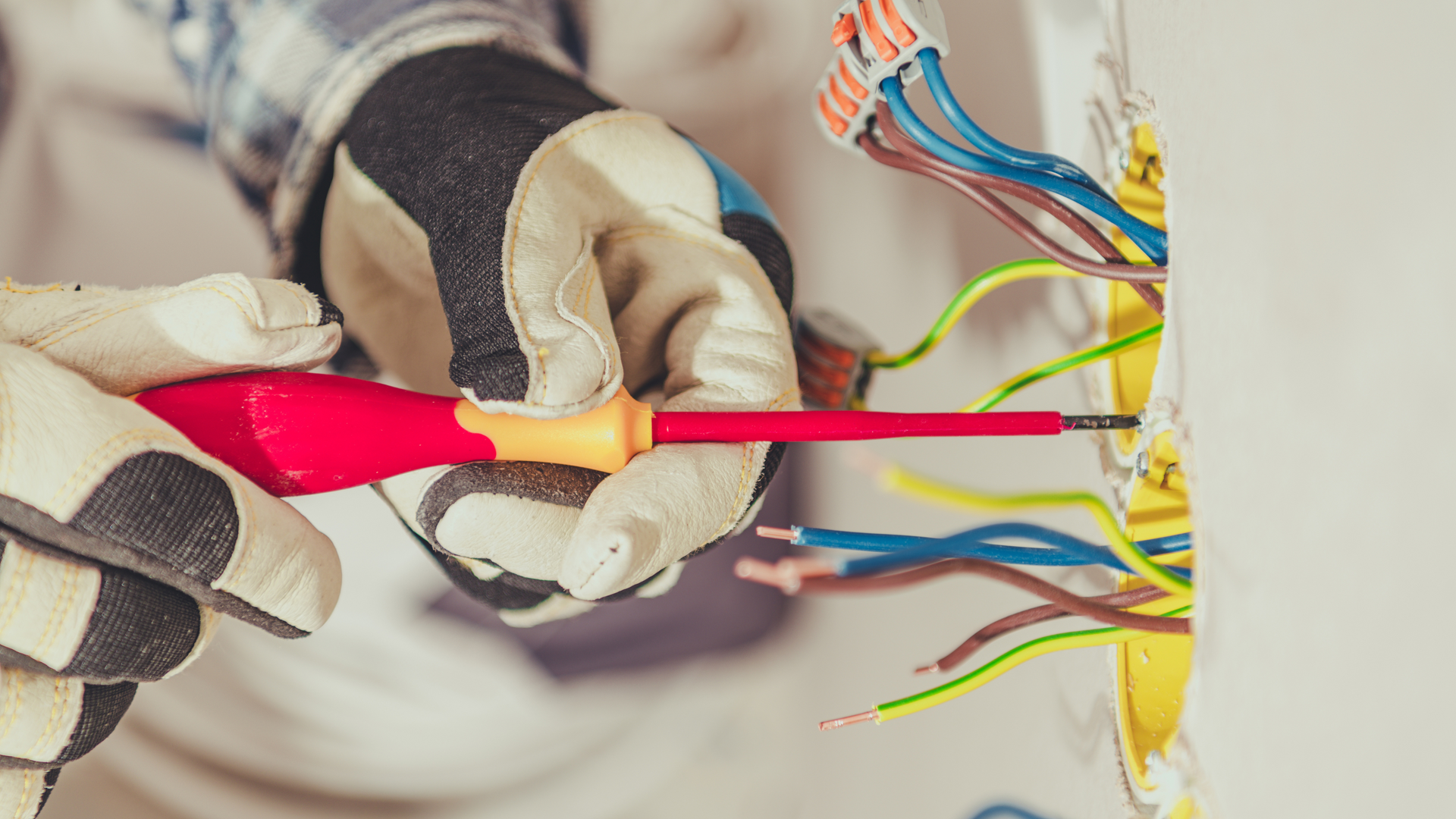 Electrician with gloves, using a red screwdriver to work on exposed wiring in a wall.