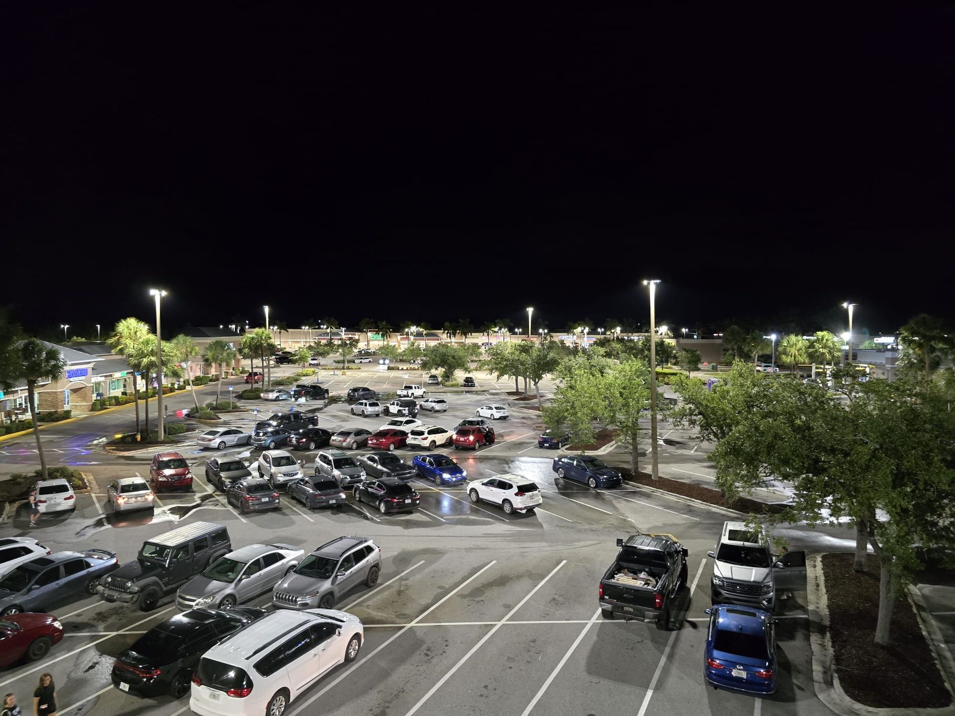 Nighttime view of a crowded parking lot under bright lights, with many parked cars and trees
