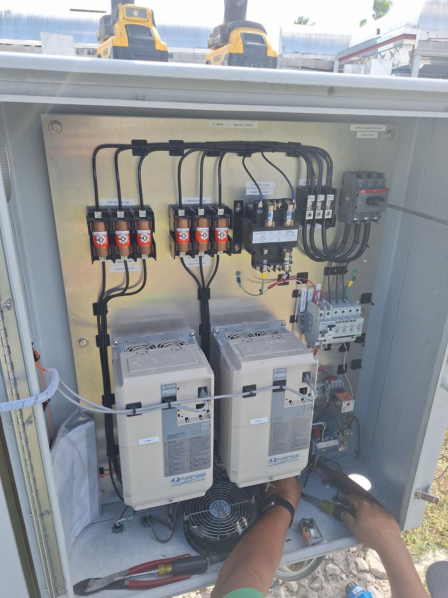 Open electrical control panel with wiring, breakers, and two drive modules; a person’s hands work inside.