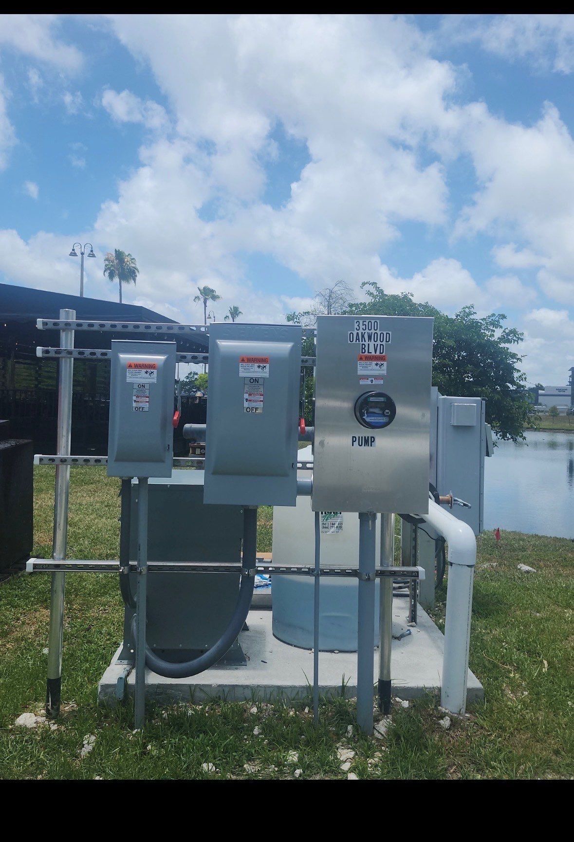 Three gray electrical control panels on a stand beside a pond under a cloudy blue sky