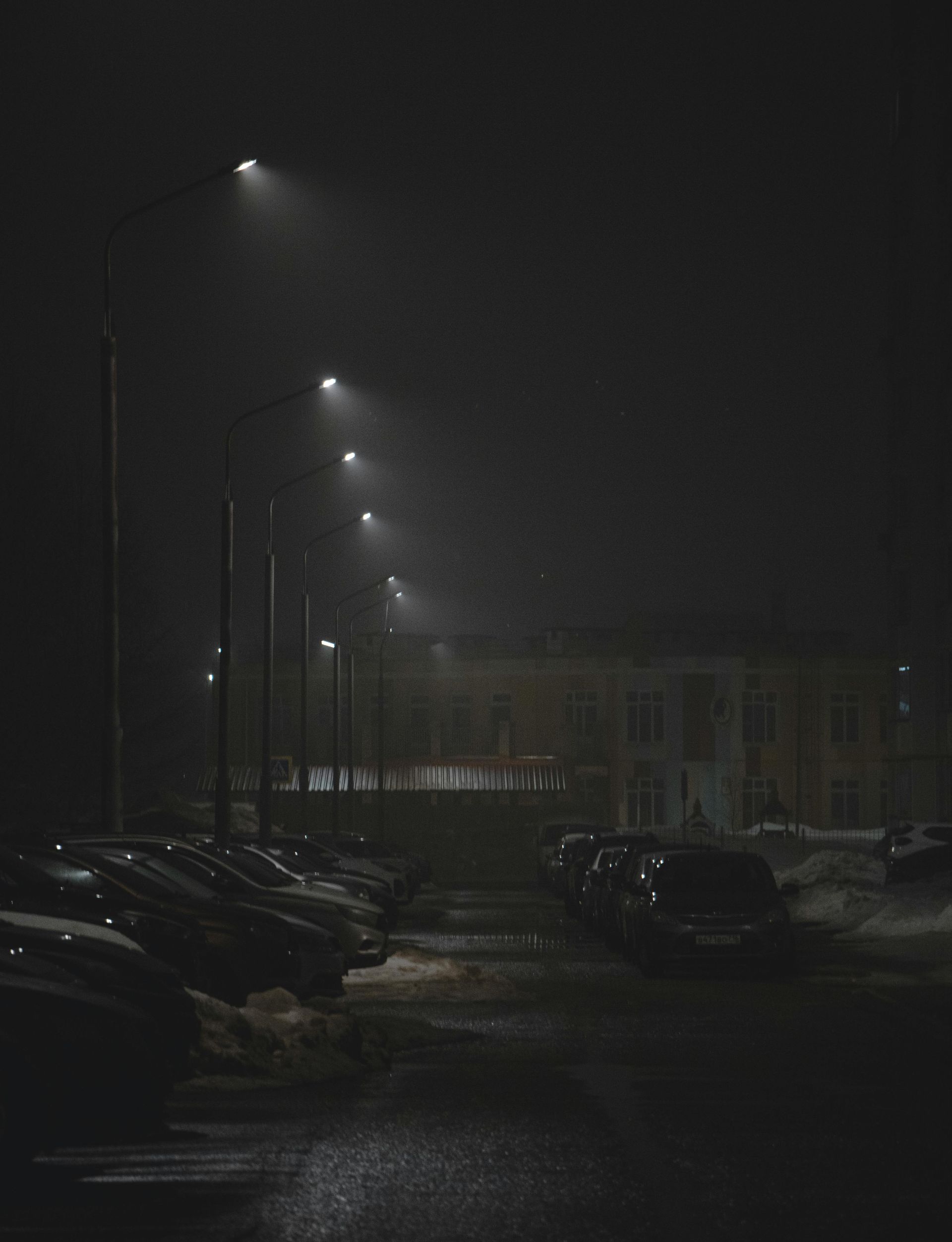A dark, nighttime parking lot with rows of cars, streetlights, and a building shrouded in fog.