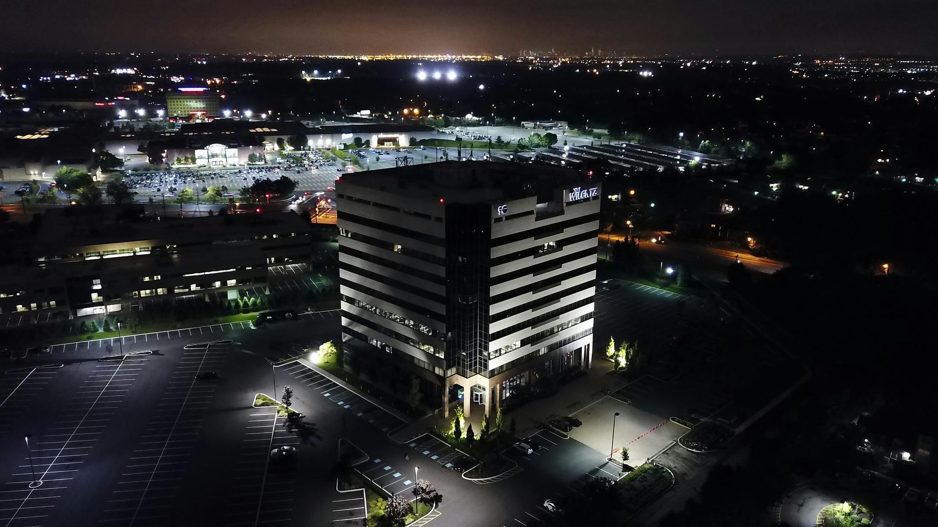 Night aerial view of a lit, multi-story office building in a parking lot, with city lights in the background.