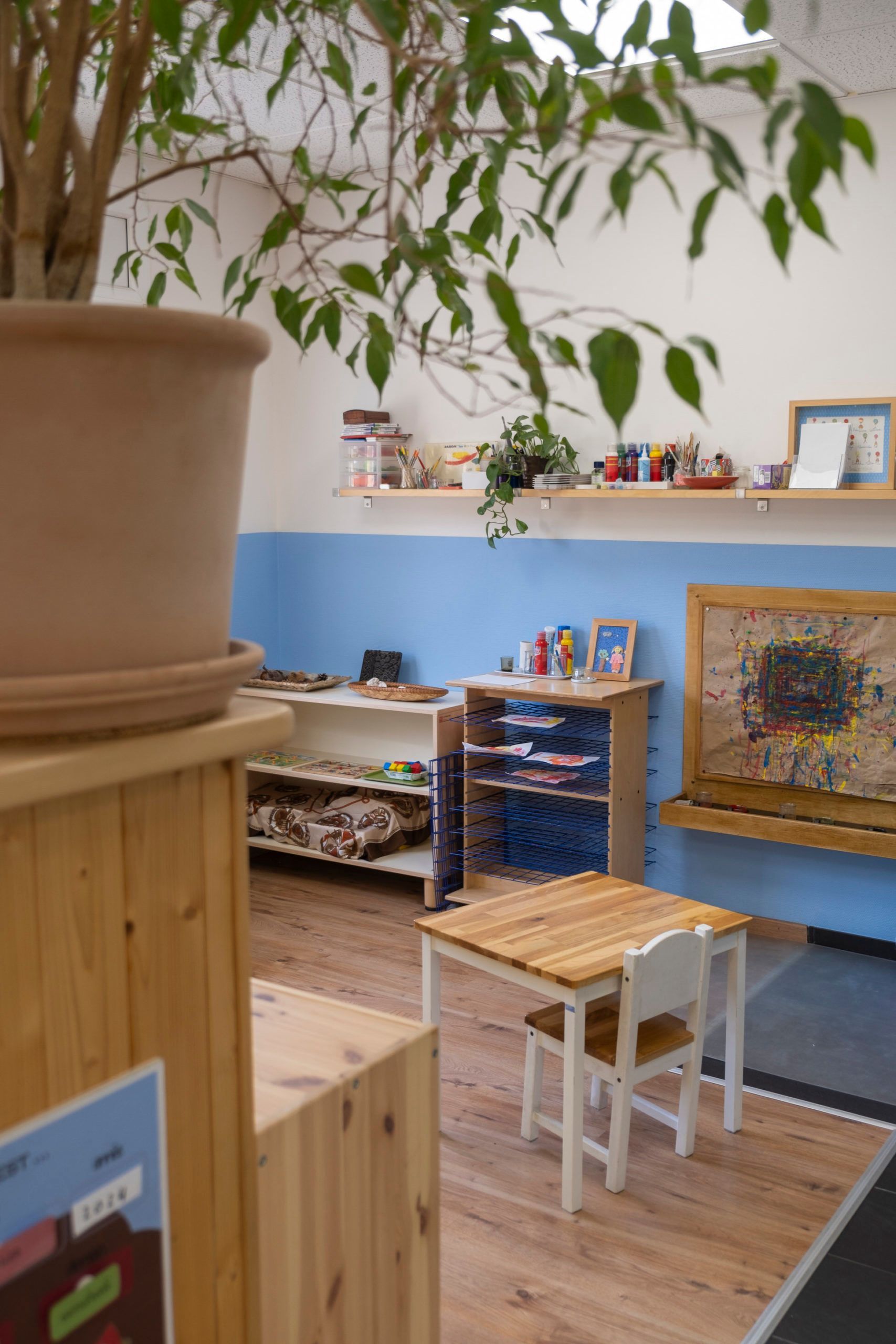 Aire de jeux confortable avec tables, chaises et étagères en bois, et une grande plante en pot au premier plan.