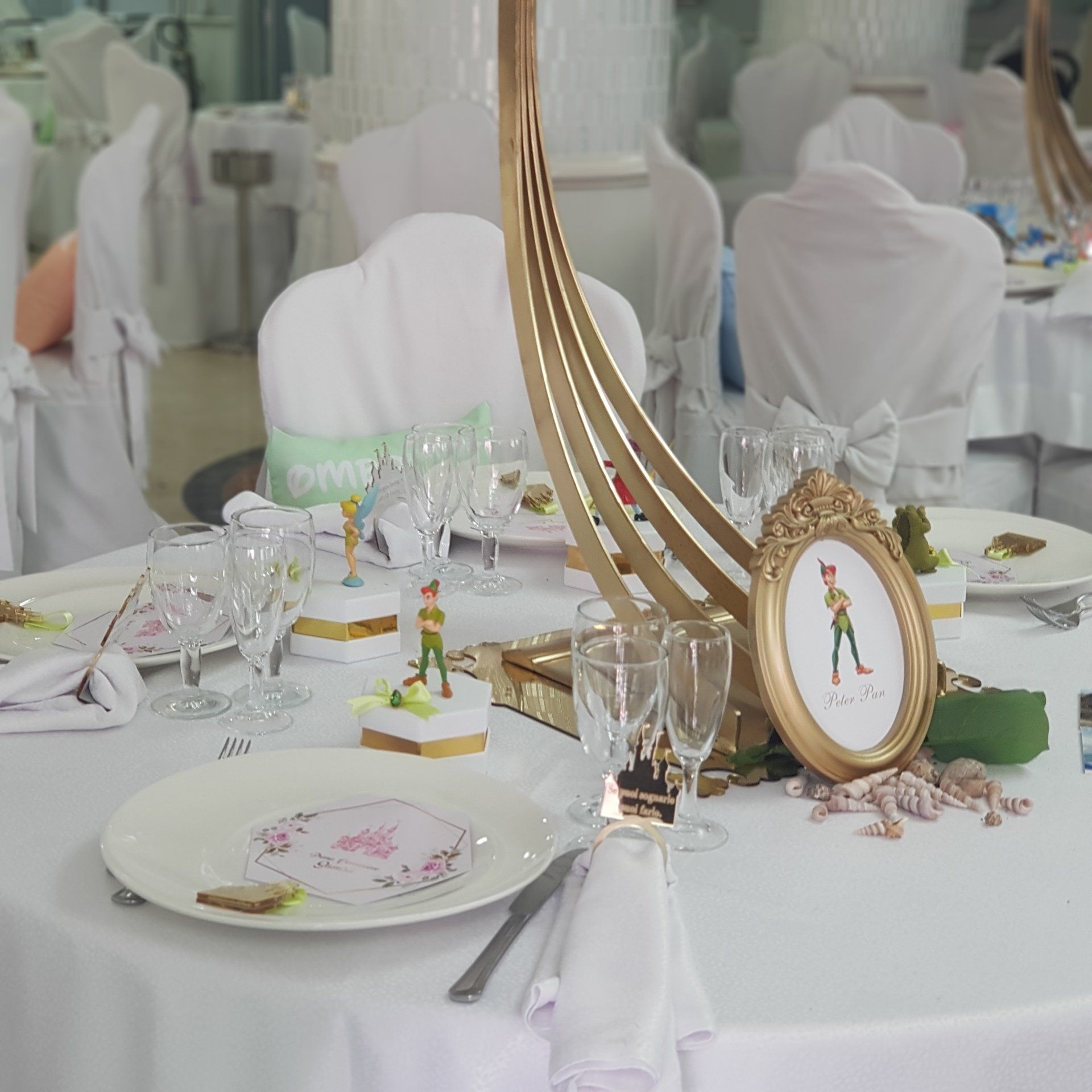 Apparecchiatura della tavola a tema Peter Pan con centrotavola dorato, opera d'arte incorniciata e tovaglia bianca.