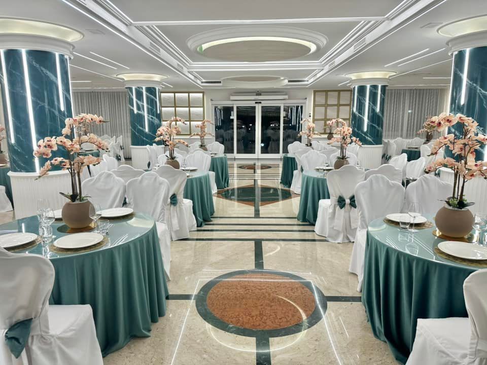 Una sala per banchetti con tavoli e sedie bianchi apparecchiati per un evento formale; illuminazione blu.