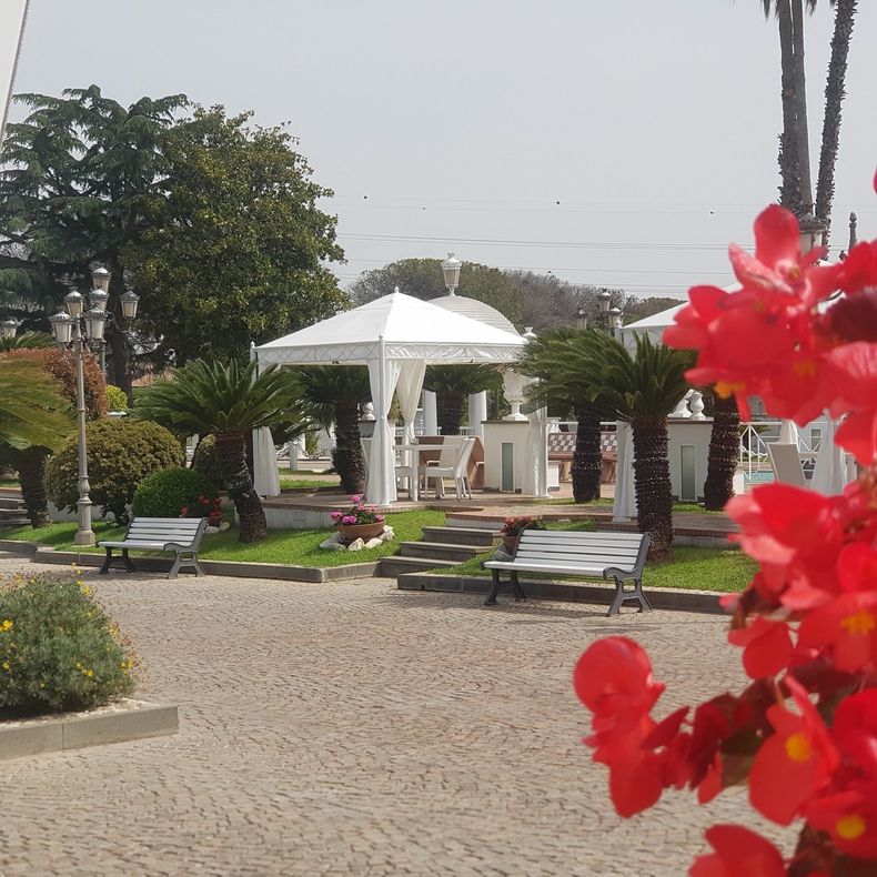 Un gazebo con tende bianche e posti a sedere in un parco con panchine, fiori e vista sul mare.