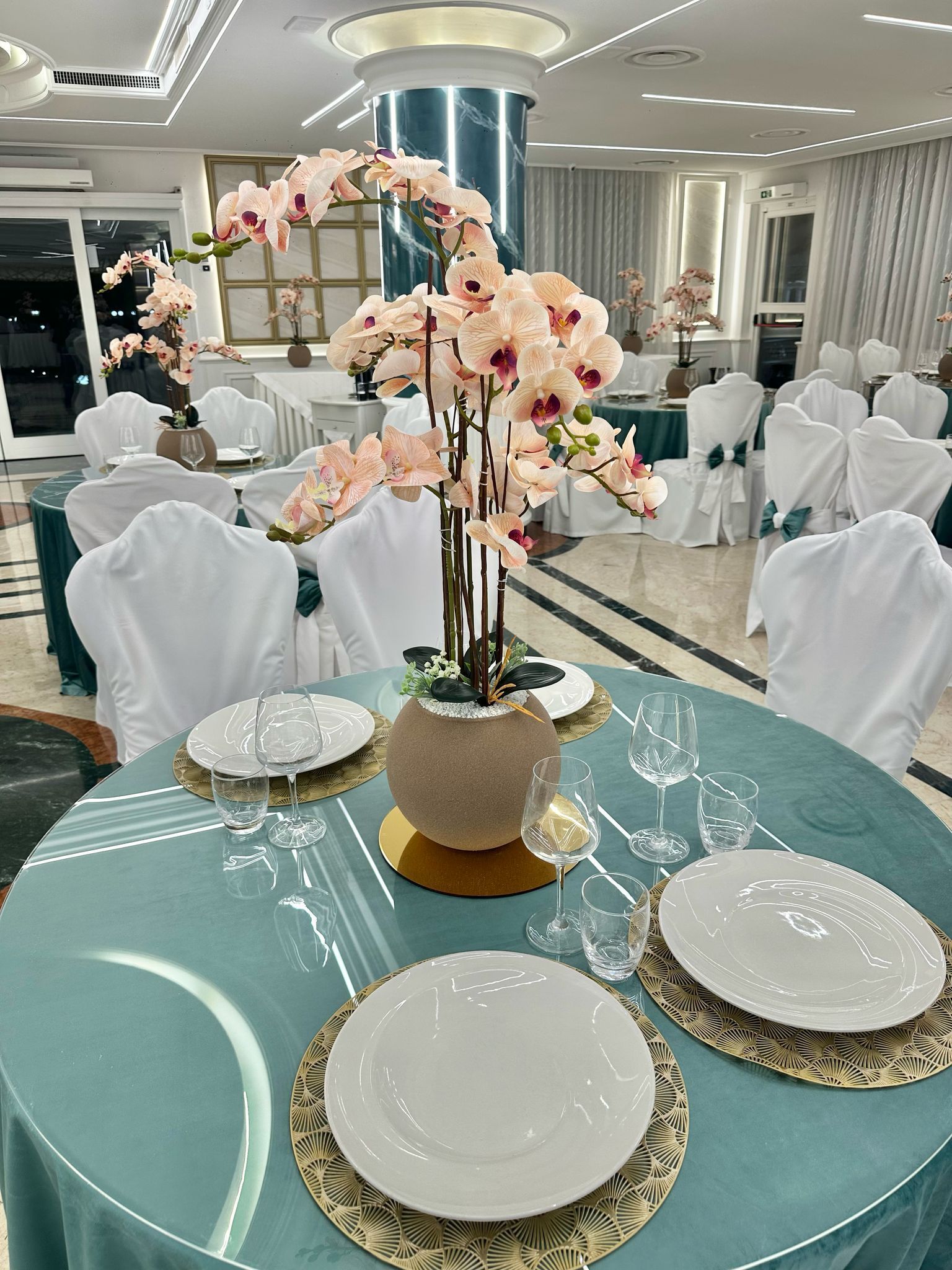 Elegante apparecchiatura con orchidee, tovaglia verde acqua, dettagli dorati e sedie bianche in una sala per banchetti.
