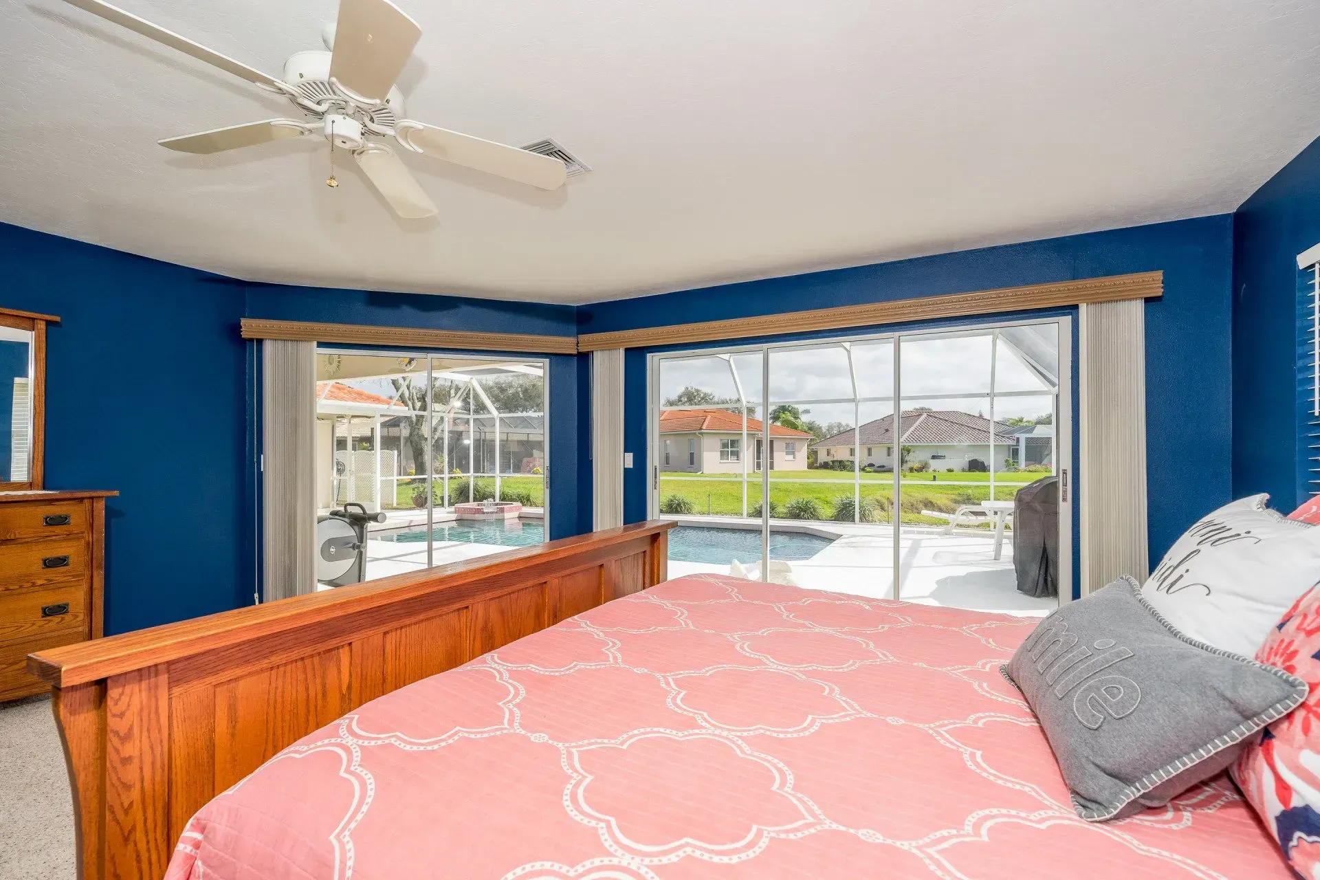 Bedroom with blue walls, wood bed and dresser, looking out onto a pool and yard through large windows.