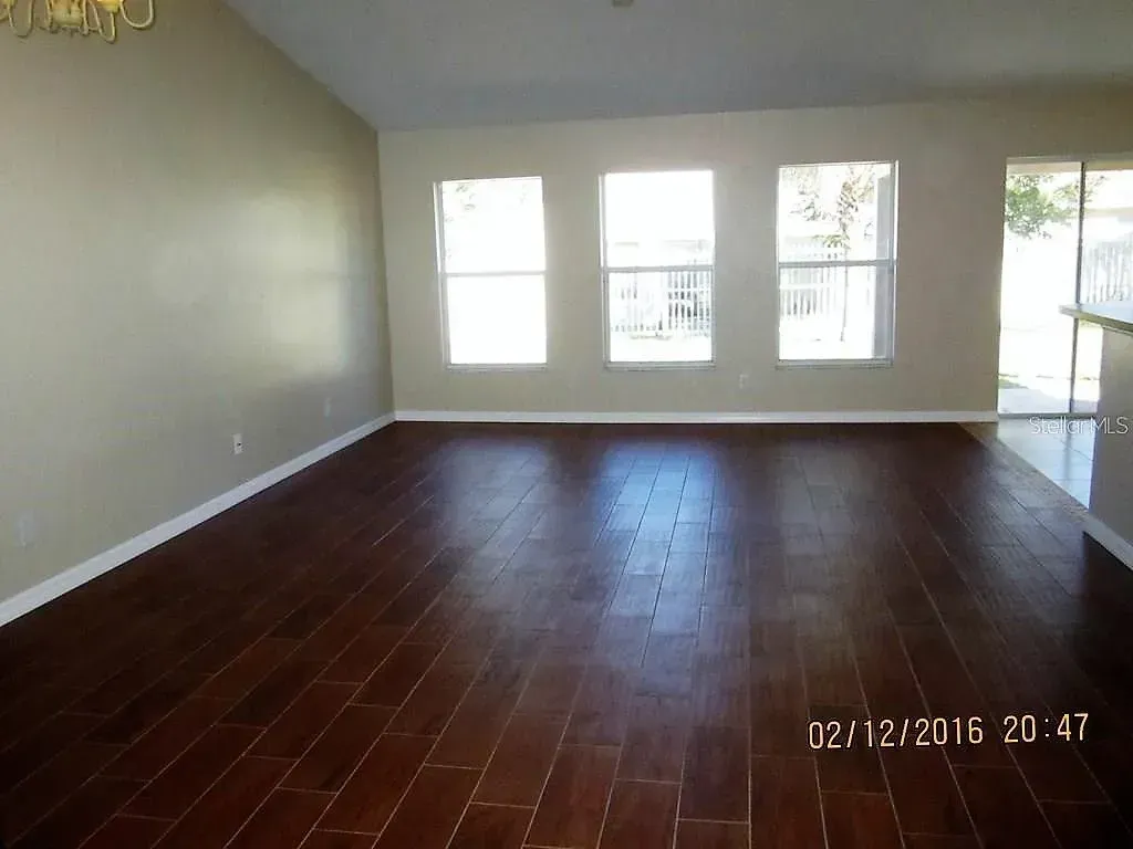 Empty living room with dark wood floor, three windows, and tan walls.