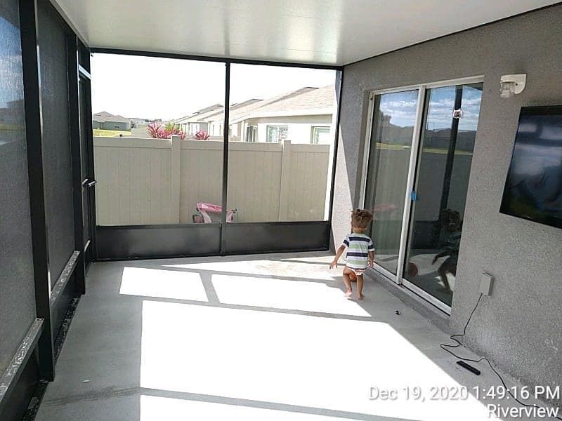 A small child walks in a screened-in porch, looking toward a sliding glass door. The floor is concrete.