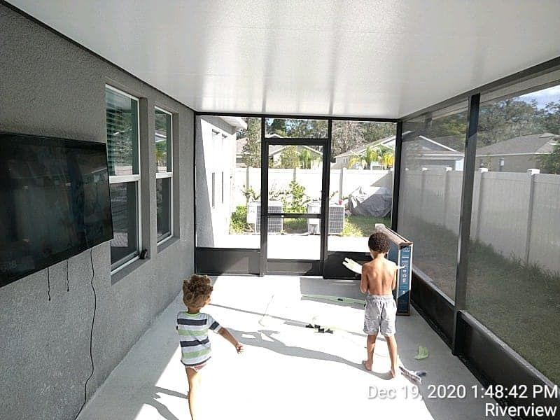 Two children play in a screened-in porch. The room has a TV on the wall, and looks out onto a yard.
